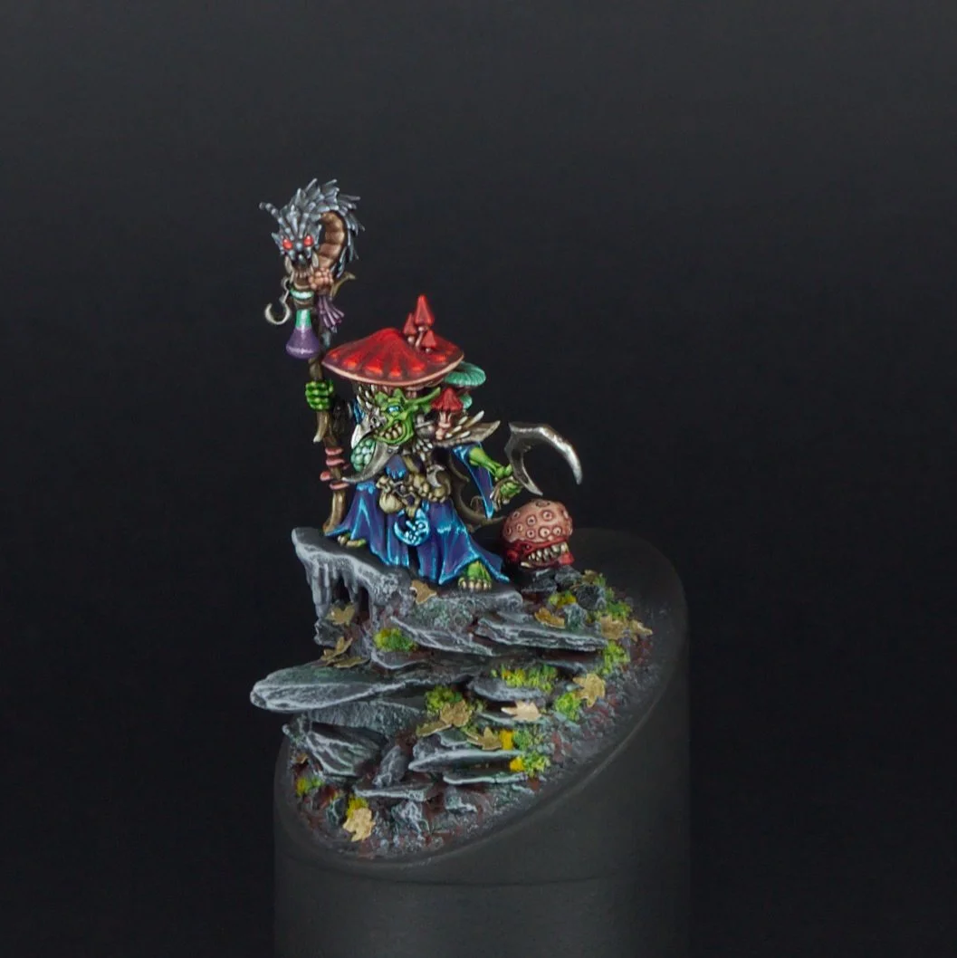 Detailed painted miniature figure of a green-skinned creature wearing a large red mushroom cap as a hat, holding a hook in one hand and a weapon in the other, with a menacing expression, standing on rocky terrain with some greenery, against a black b