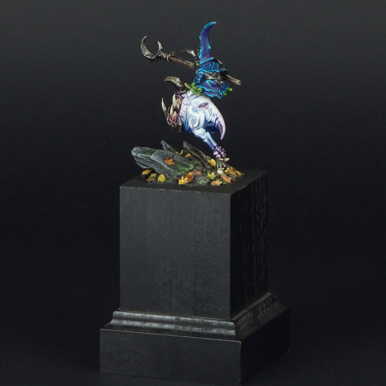 A miniature figurine of a fantasy mage character in a vibrant blue robe and pointed hood, wielding a staff with a crescent-shaped top, stands on a detailed base with rocks and autumn leaves, displayed on a black pedestal against a black background.