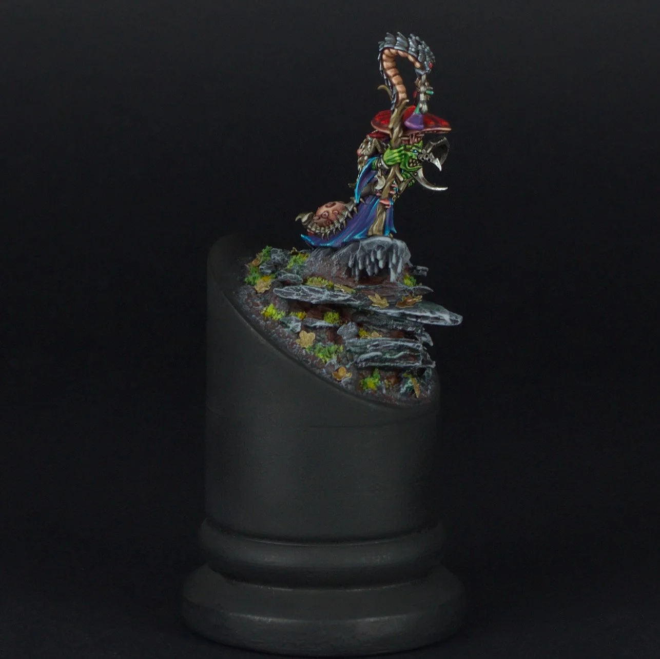 Colorful miniature figure of a dragon perched on rocks with foliage, displayed on a black stand against a dark background.