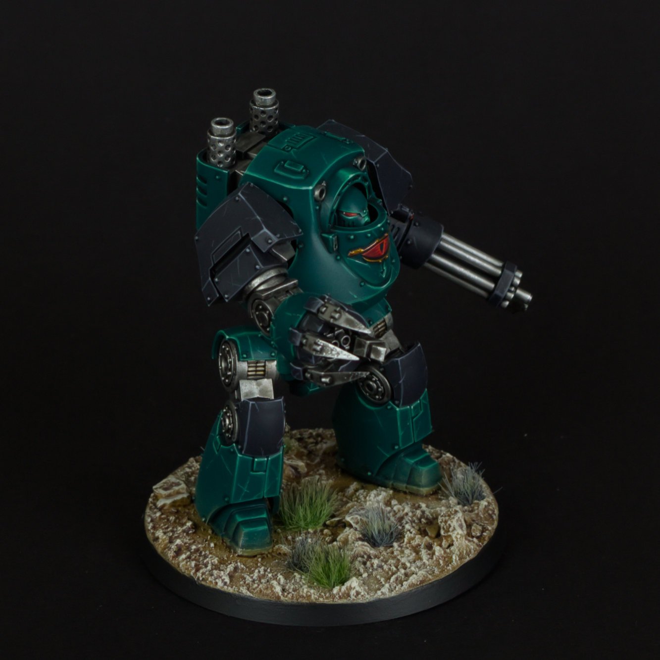 A painted miniature model of a futuristic teal robot with heavy armor, weapons, and a mining helmet, standing on a textured base with small rocks and grass on a black background.