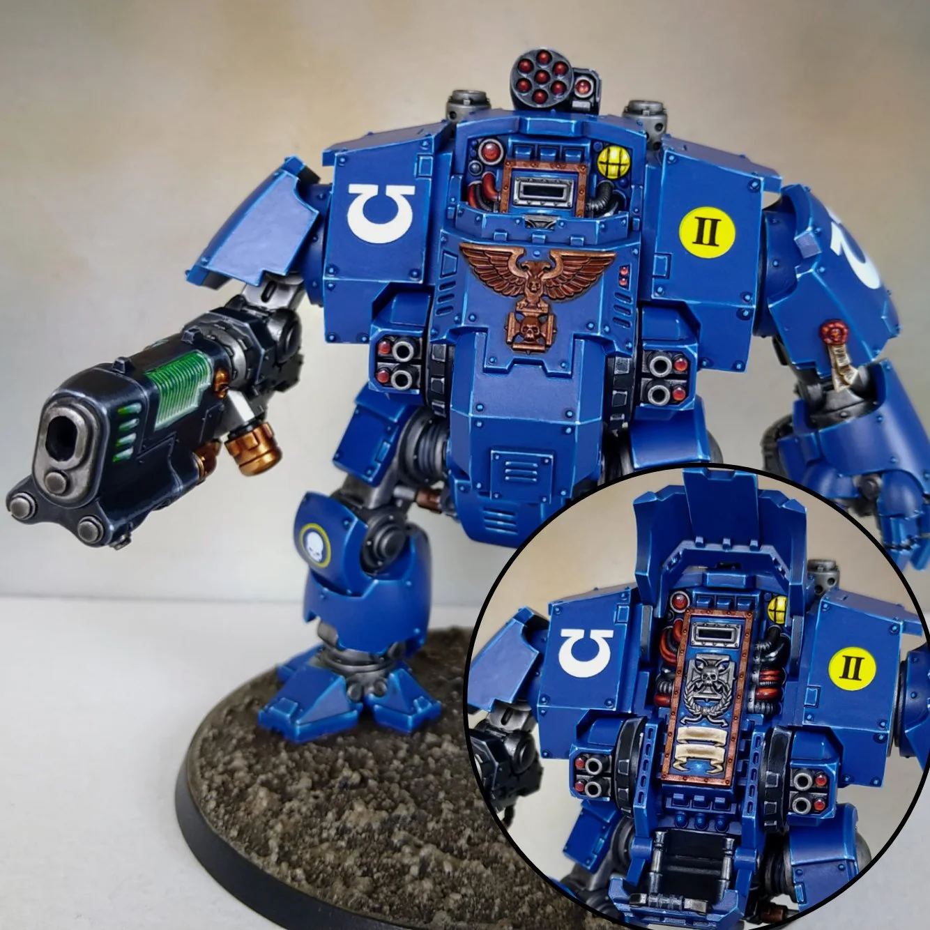 A detailed model of a blue armored sci-fi robot figure with weaponry, standing on a textured circular base. The robot has a golden eagle emblem on its chest, various caution markings, and exposed mechanical parts, with a close-up inset showing the ro