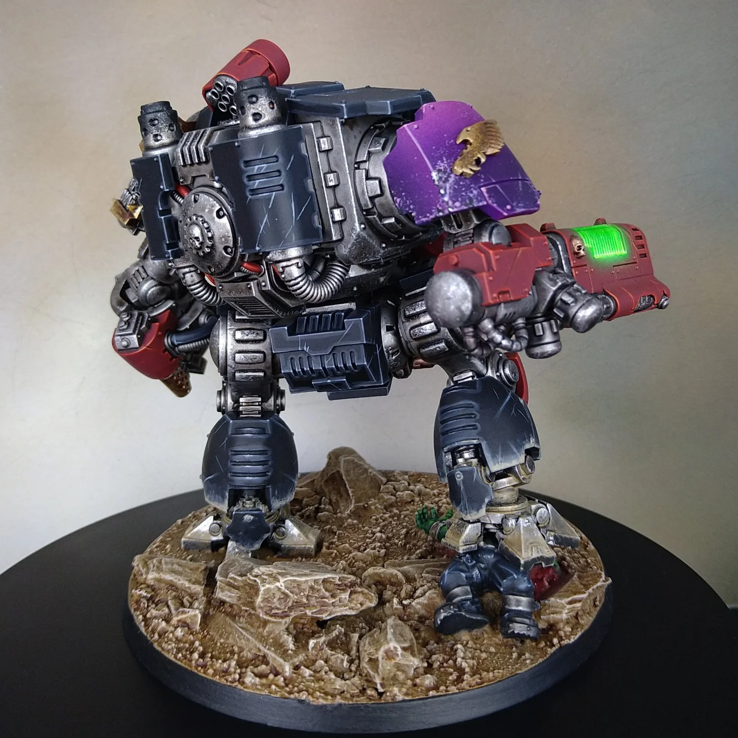 A detailed miniature model of a robotic war machine with a large purple armor plate and a weapon, standing on a rocky diorama base.