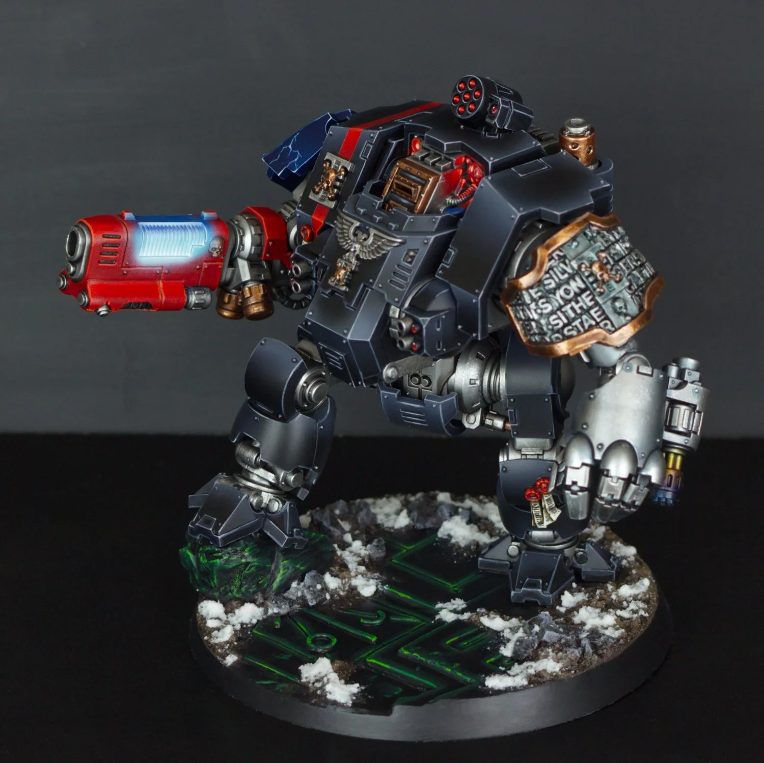 A detailed miniature model of a war machine or robot with black, red, and metallic gold and silver details, standing on a snow-textured base with green glowing accents.