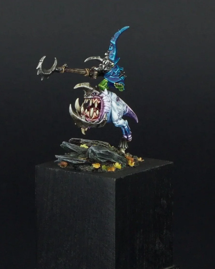 A painted miniature figurine of a goblin riding a monster, wielding a spear, on a rocky base with fallen leaves against a black background.
