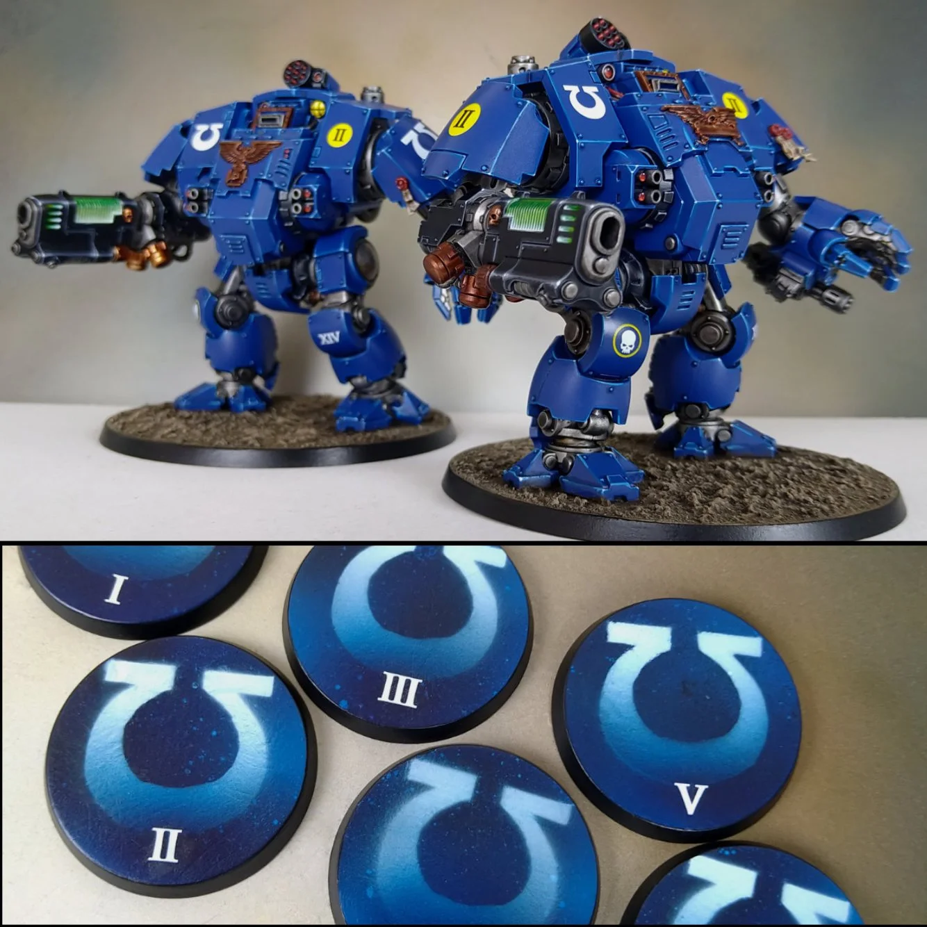 Two detailed blue tabletop miniatures of war machines on round bases, with various painted symbols and markings, along with circular tokens featuring the Imperial Aquila symbol and Roman numerals.