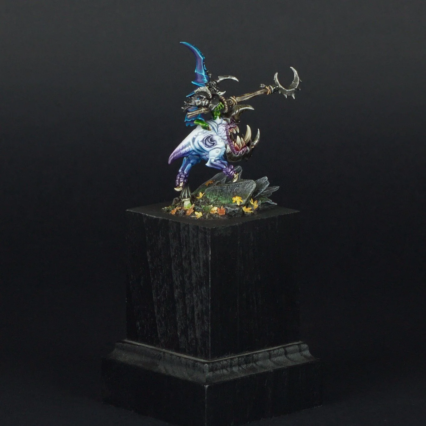 A painted miniature figure of a monstrous dragon with a rider wielding a spear, mounted on a black pedestal with autumn leaves and rocks, set against a dark background.