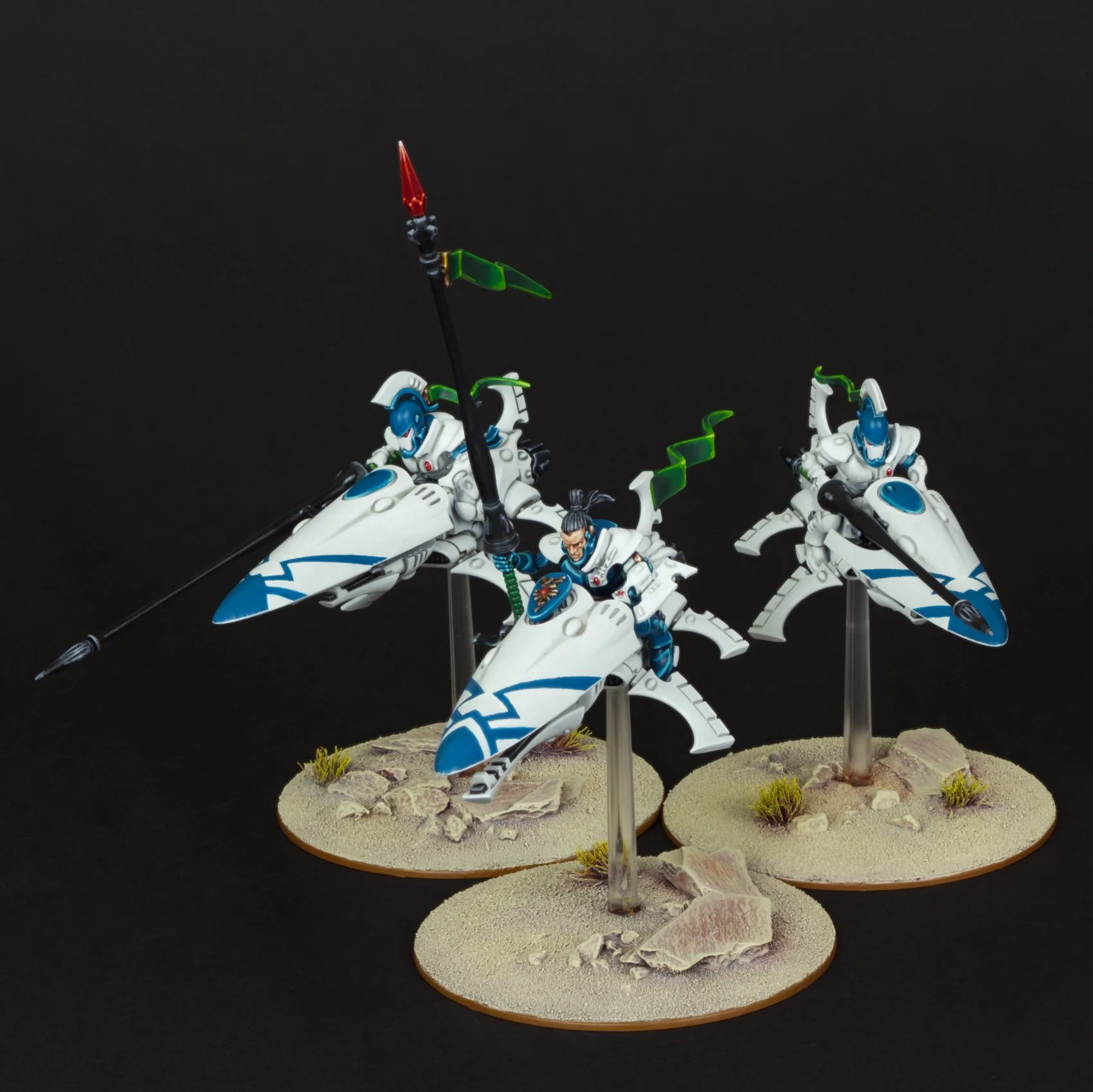 Group of three painted miniature sci-fi figures on round bases, depicting characters in white armor riding futuristic hover bikes with blue and green accents on a desert-like terrain with rocks and small plants, against a black background.