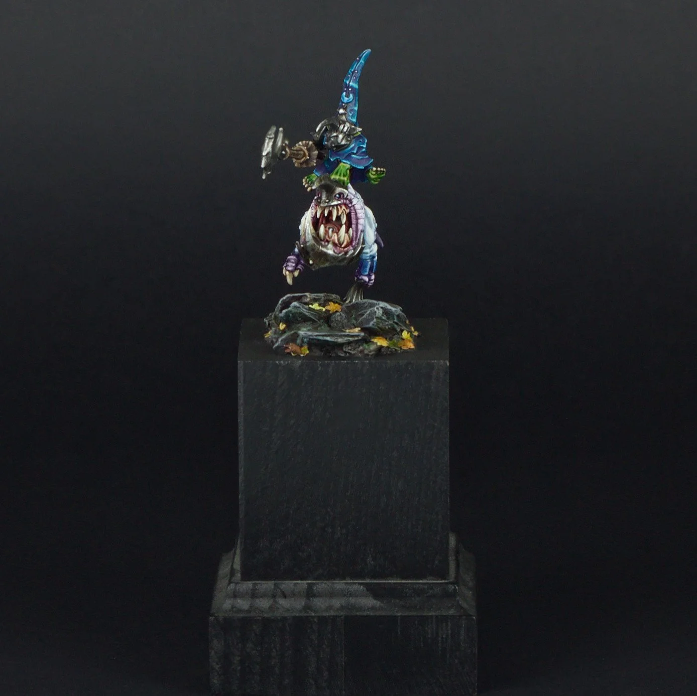 A miniature figurine of a fantasy warrior riding a monstrous creature, mounted on a textured base with autumn leaves, displayed on a black pedestal against a black background.