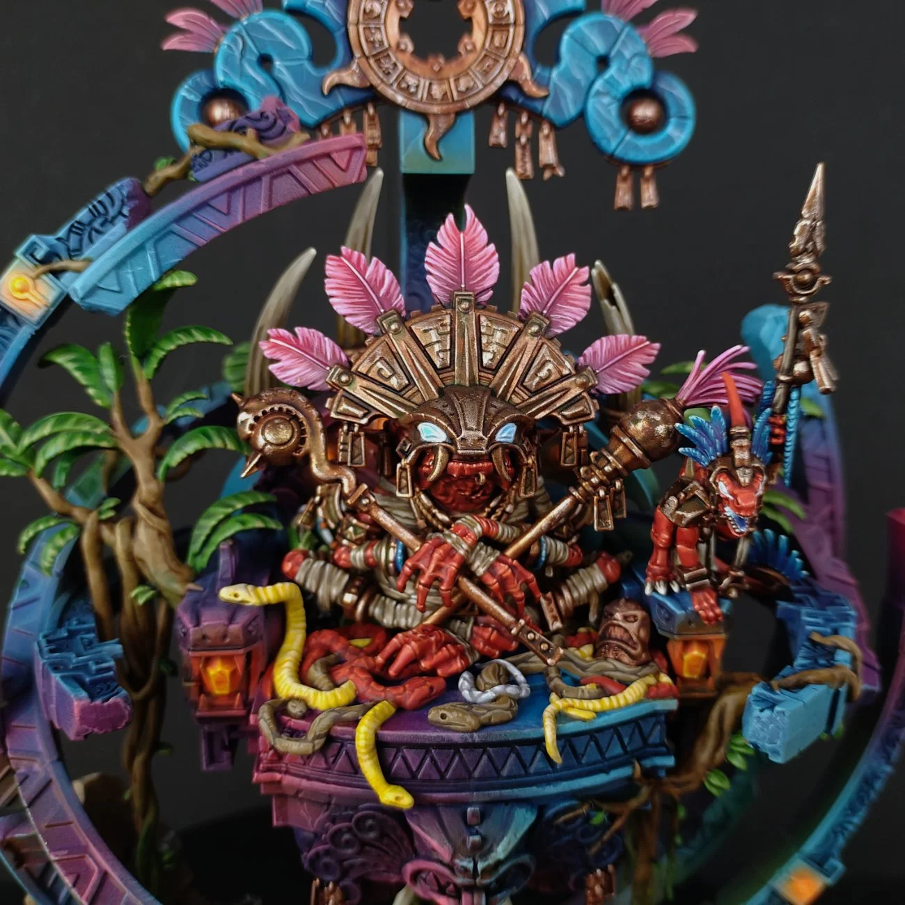 Colorful figurine depicting a tribal warrior with pink feathers, holding a spear, sitting on a throne surrounded by jungle leaves, snakes, and other ornate details.