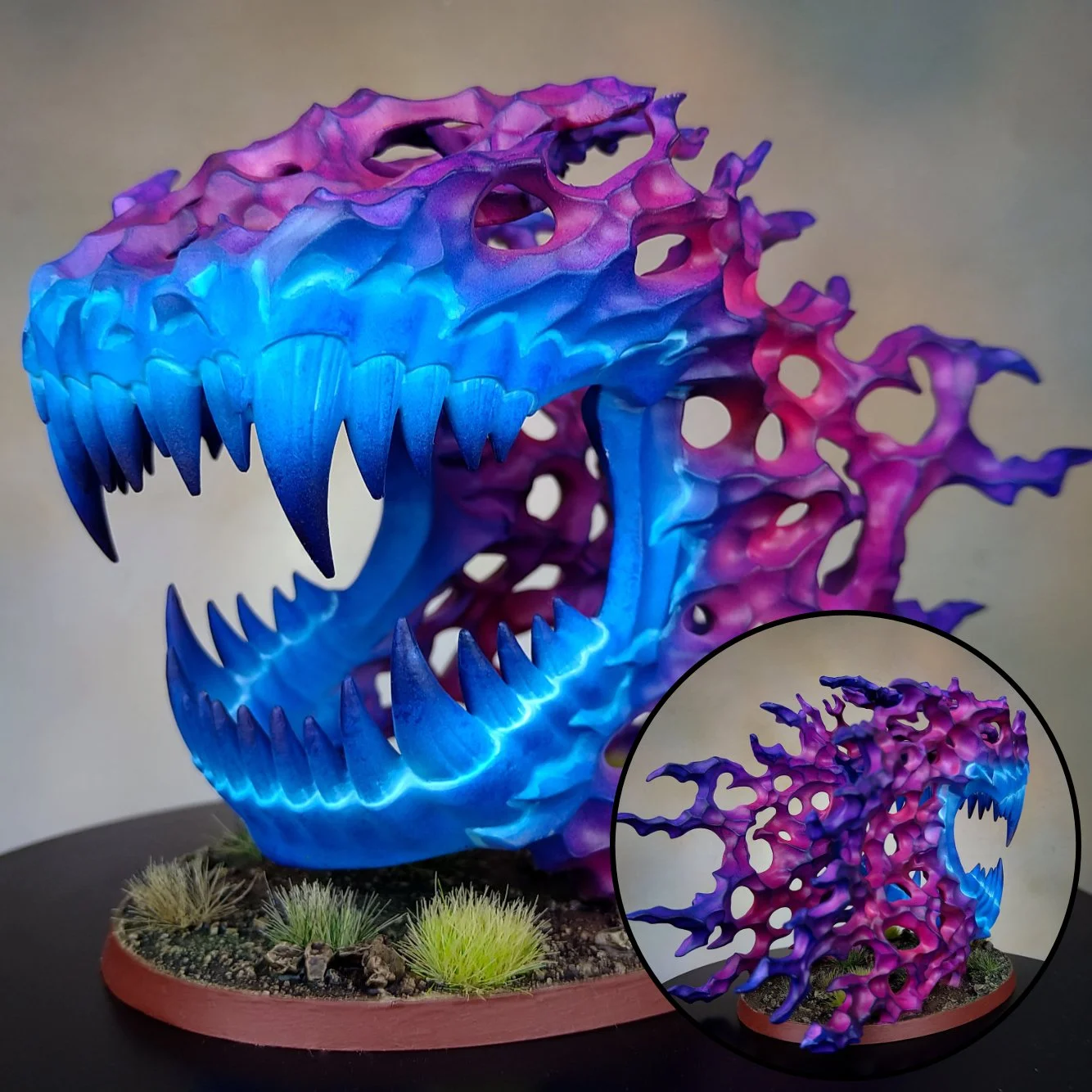 Colorful sculpture of a dinosaur skull with large sharp teeth, made with an intricate, lacy, coral-like structure in shades of blue, purple, and pink, displayed on a circular base with small artificial plants.