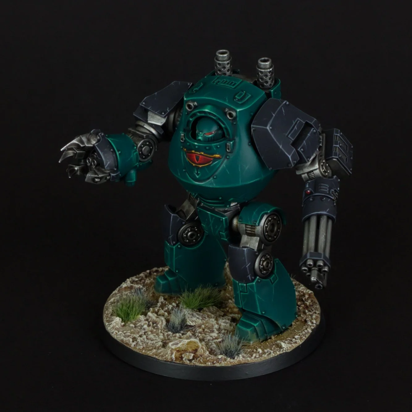 A detailed model of a teal-colored robot with large arms, tank-like legs, and weapon attachments, standing on a round base with rocks and grass, against a black background.