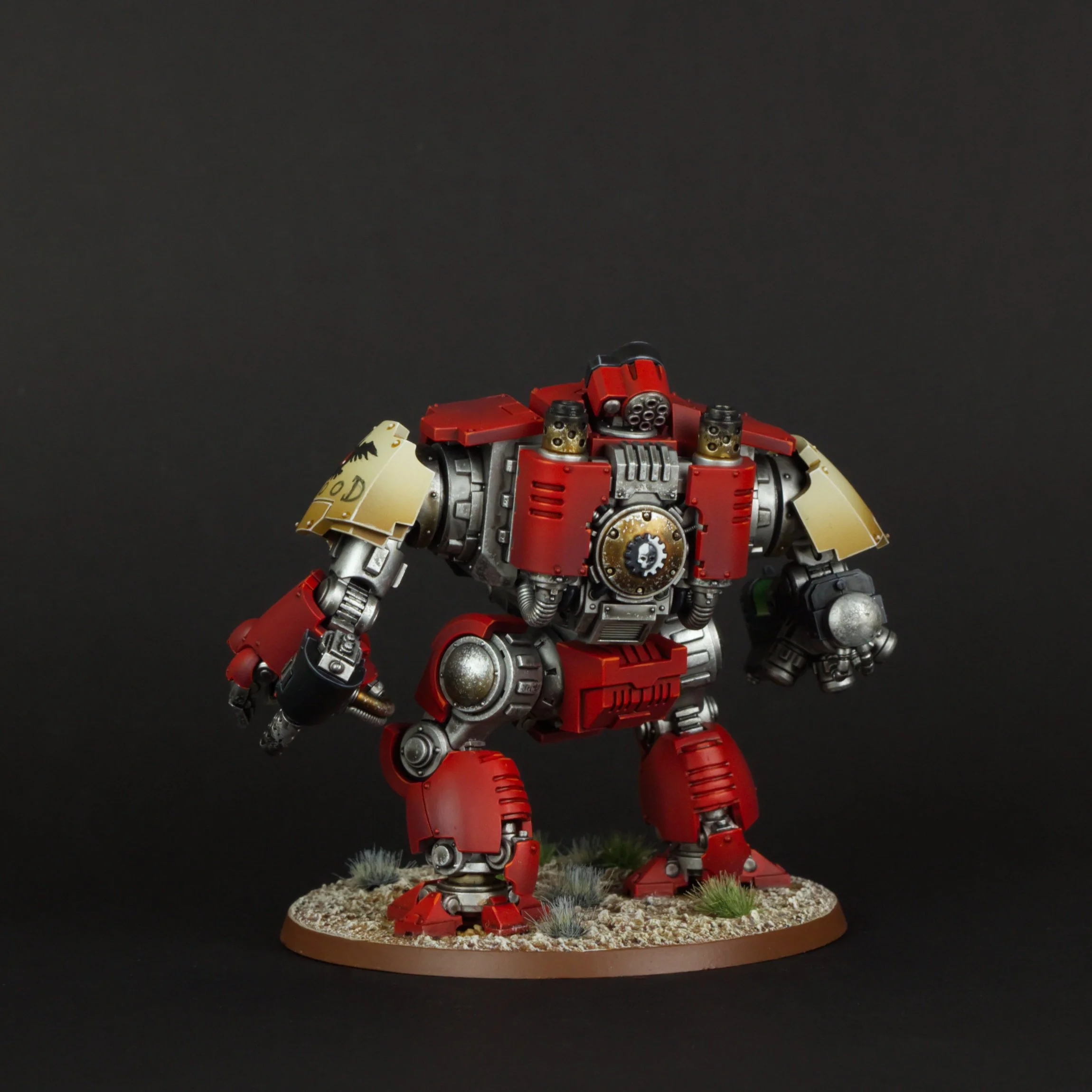 A detailed miniature sculpture of a red and silver mecha robot standing on a round base decorated with small rocks and tufts of grass against a black background.