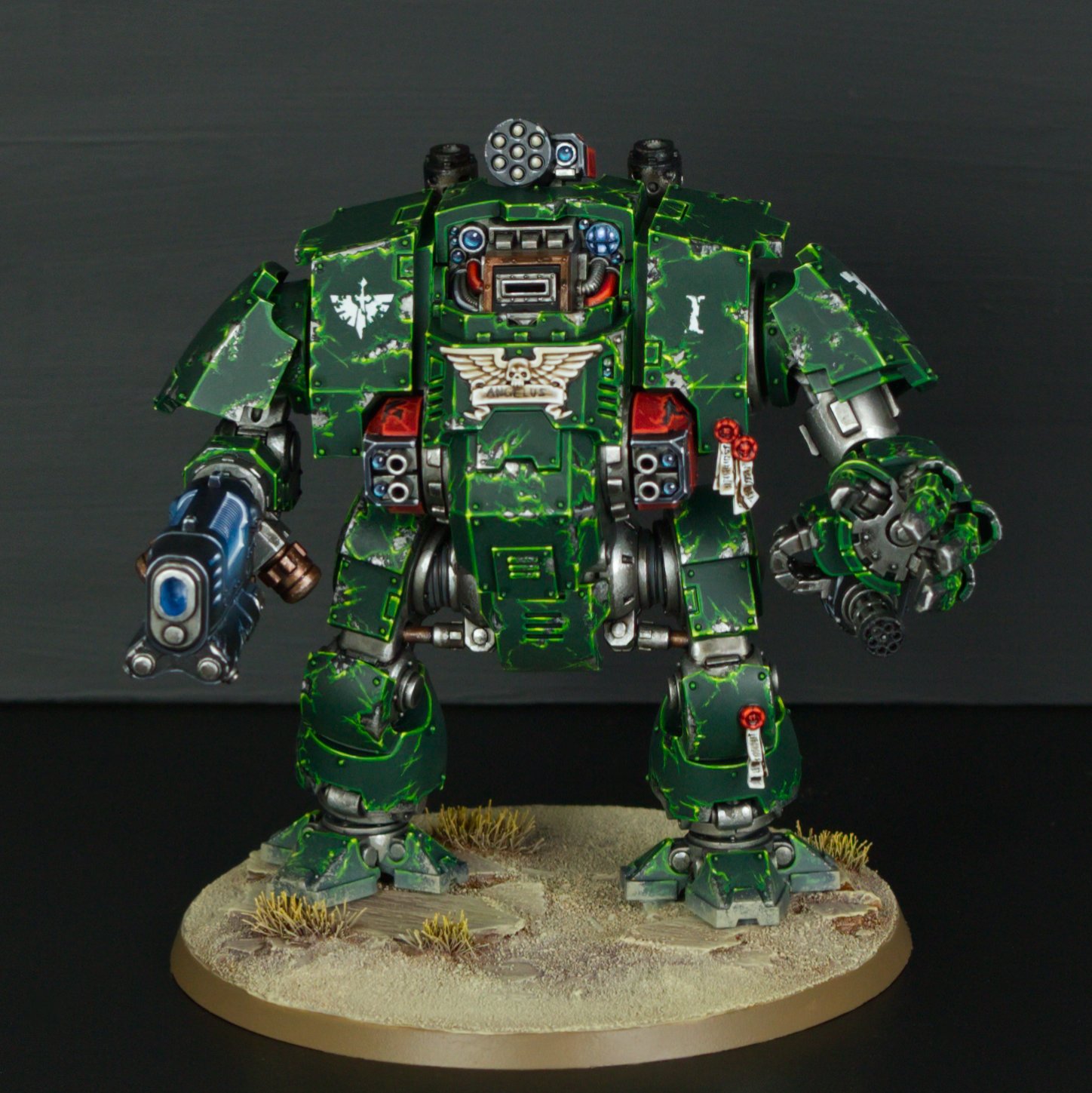 A model of a green armored mech with black and silver details, standing on a desert-like base with small tufts of grass, against a black background.