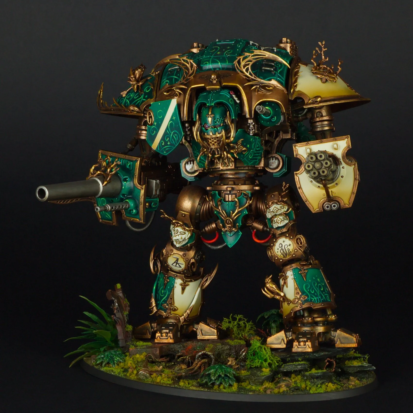 A highly detailed miniature model of a large, green and gold armored robot with skull and dragon motifs, standing on a base with grass and plants, against a black background.