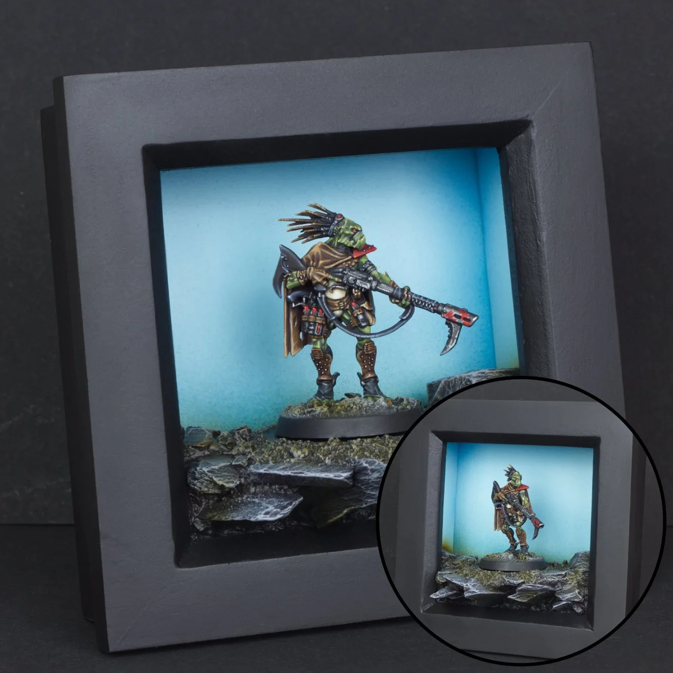 Miniature figurine of a green alien or orc-like creature dressed in armor and a brown cape, holding a weapon with a red blade, displayed inside a black shadow box with a blue background and rocky terrain at the base.