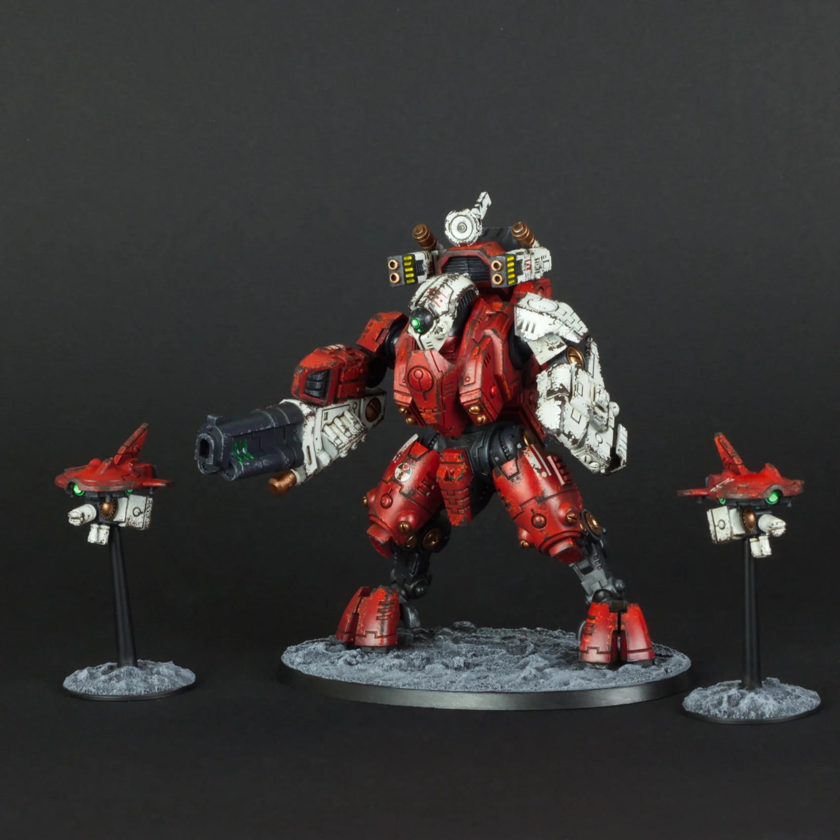 A detailed model of a red and white futuristic robot with black accents, standing on a rocky base against a black background. The robot has multiple weapons and attachments, with two smaller robotic figures on either side, also mounted on rocky bases