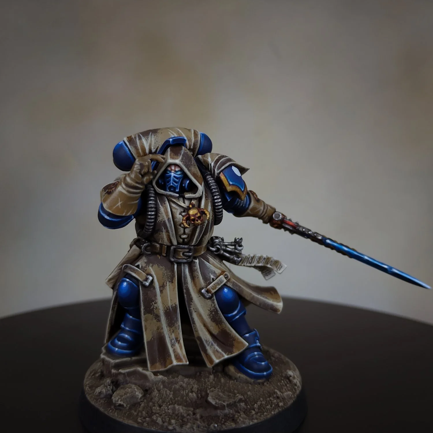 A painted miniature figurine of a sci-fi warrior in blue and beige armor, holding a blue sword and pointing with his right hand, standing on a textured base.
