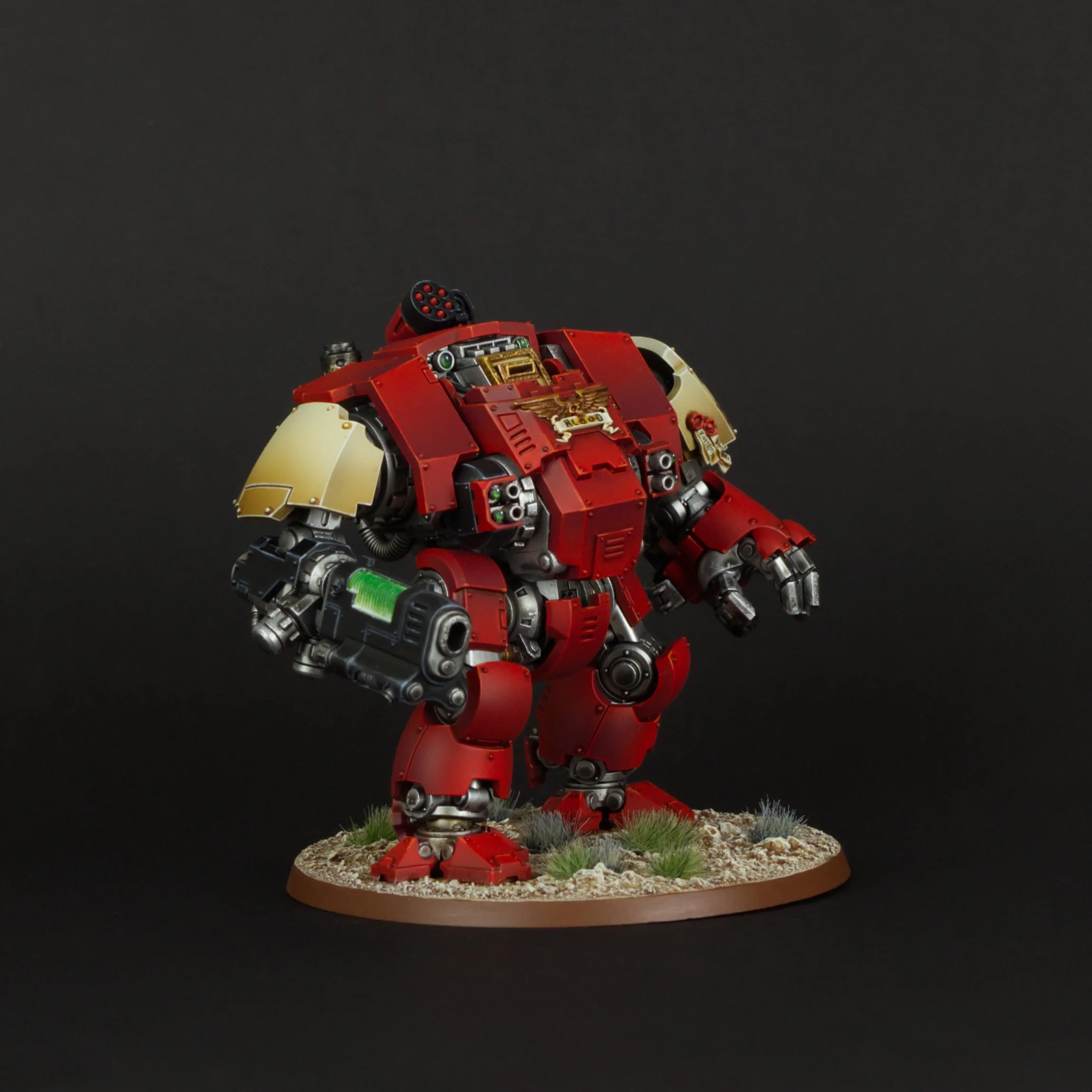 A detailed model of a Warhammer 40k red and gold robot standing on a round base with small plants and rocks, against a black background.