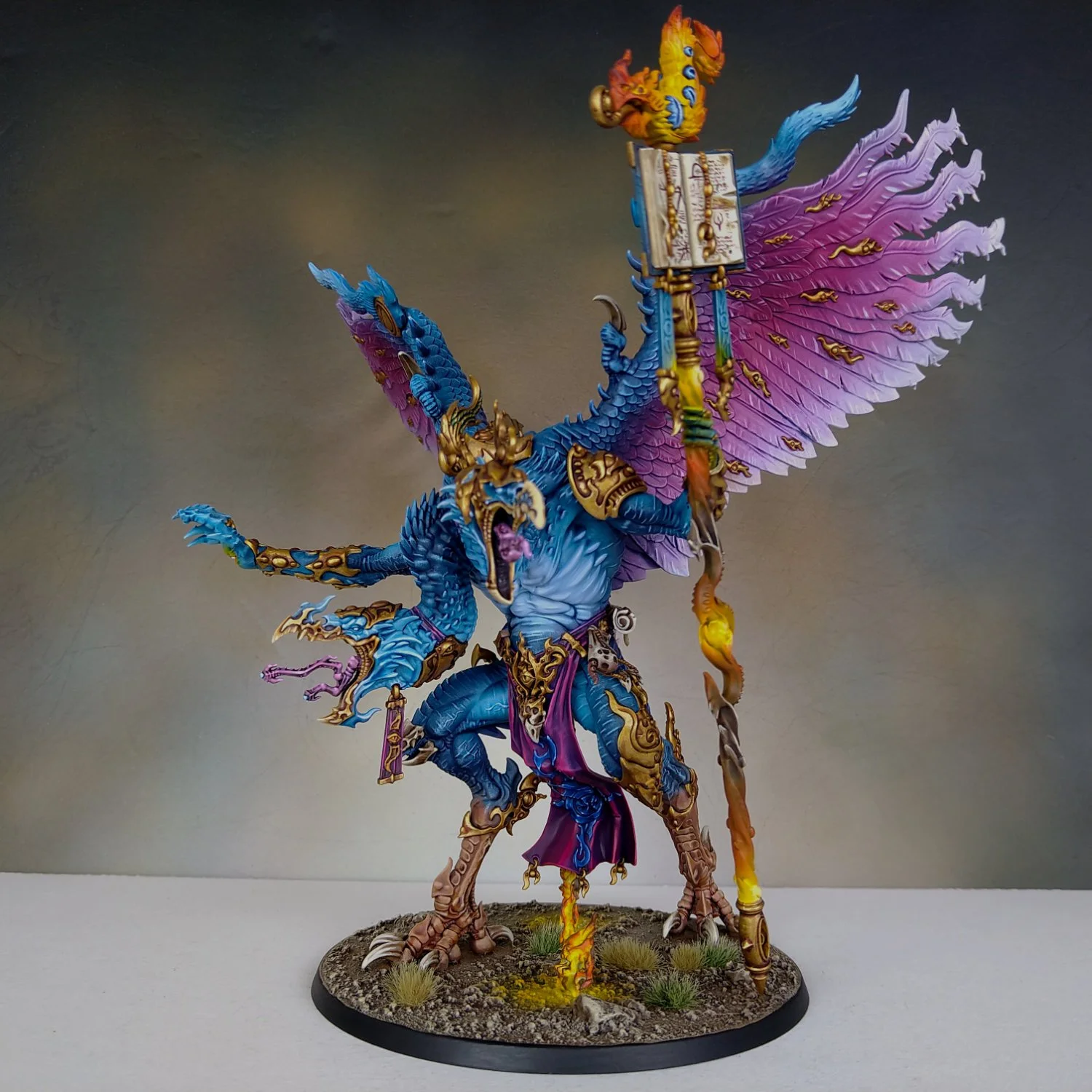 A highly detailed miniature figure of a mythical dragon-like creature with vibrant blue, purple, and gold armor, large pink wings, and an open mouth showing sharp teeth, mounted on a textured round base with grass and rocks.