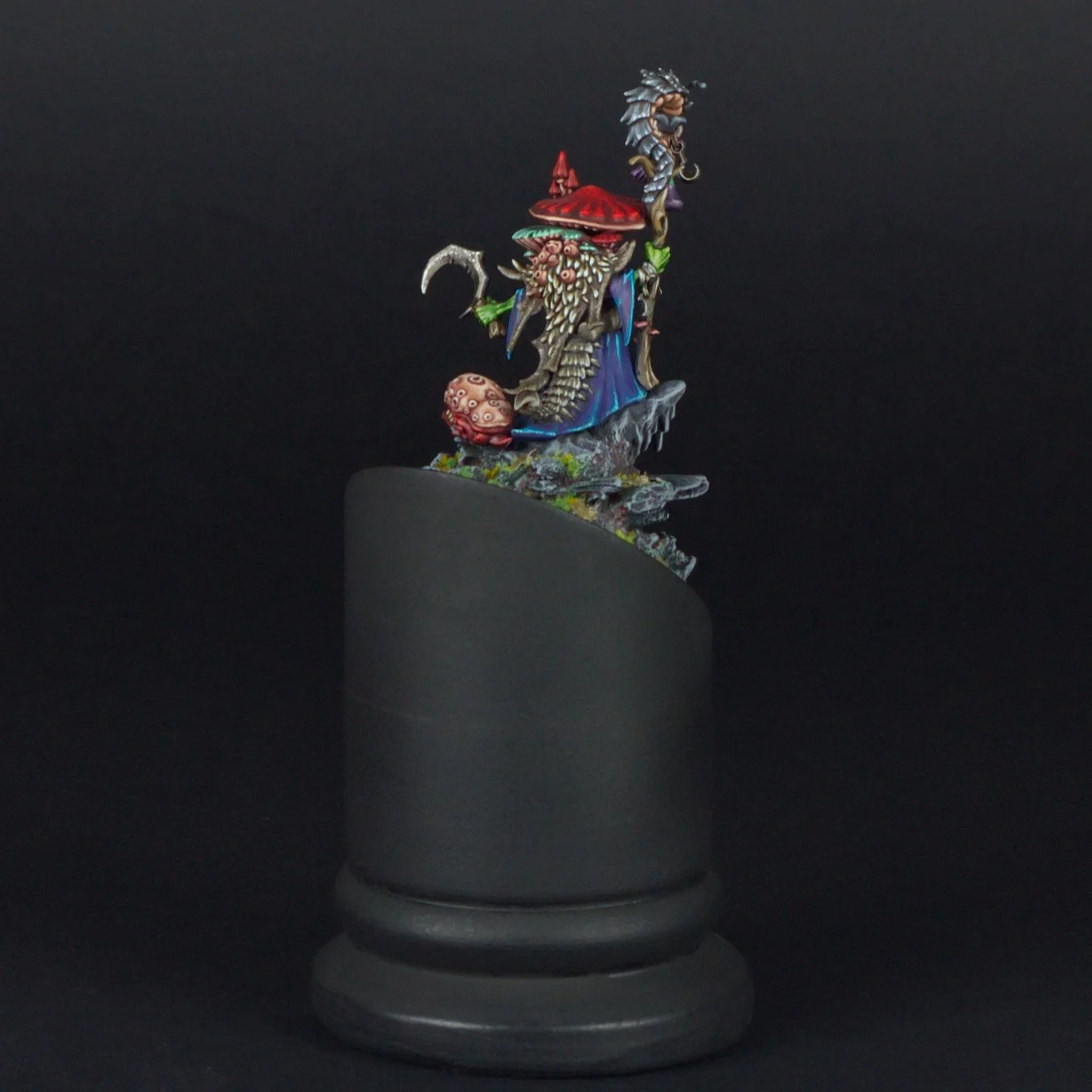 Colorful fantasy figurine of a wizard with a long beard, holding a scythe, wearing a red pointed hat, standing on a rocky base, displayed on a black cylindrical pedestal.