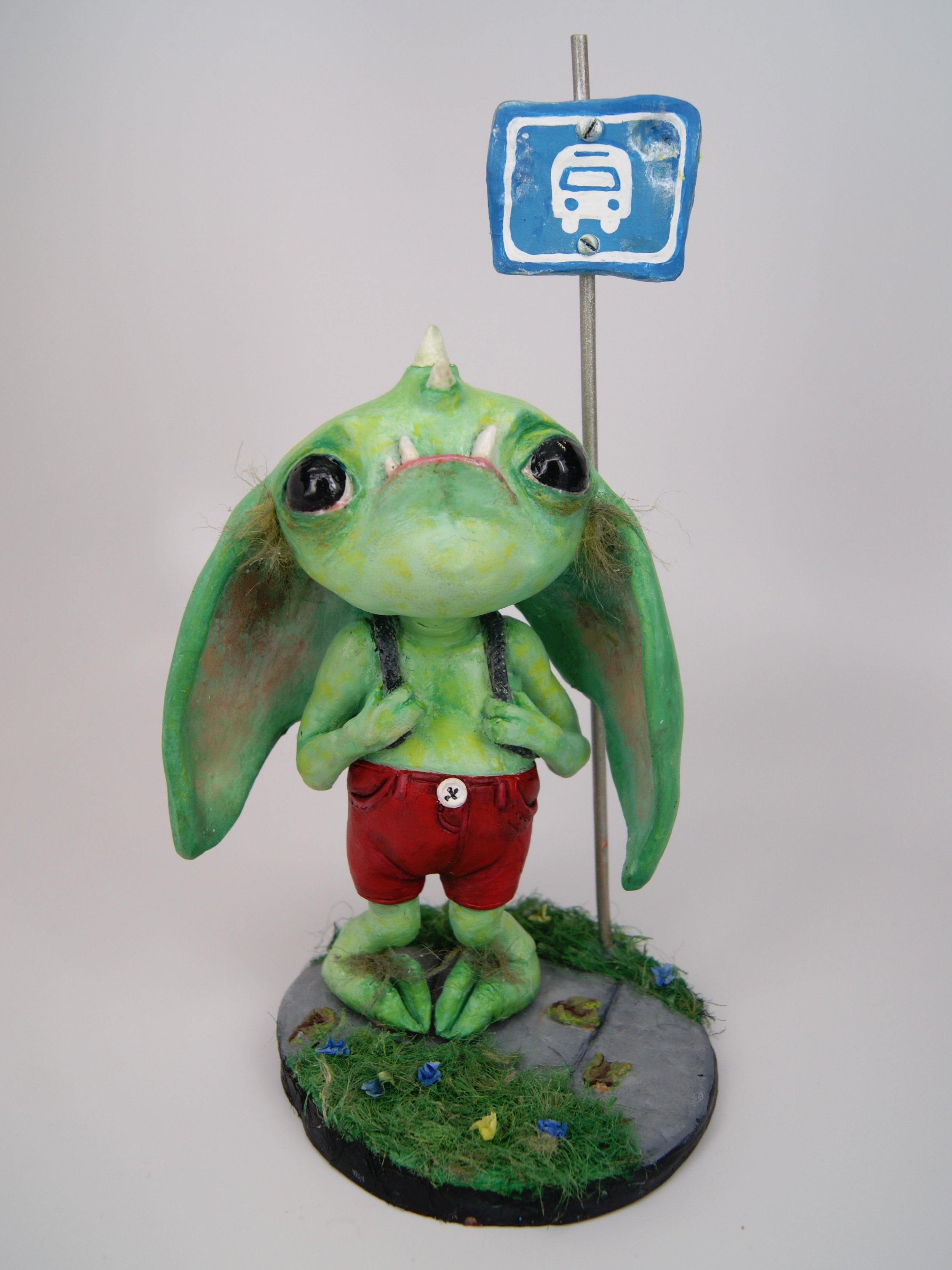 A sculpture of a green, anthropomorphic creature with large eyes, wearing red shorts, standing on a grassy base with a sidewalk, near a bus stop sign.