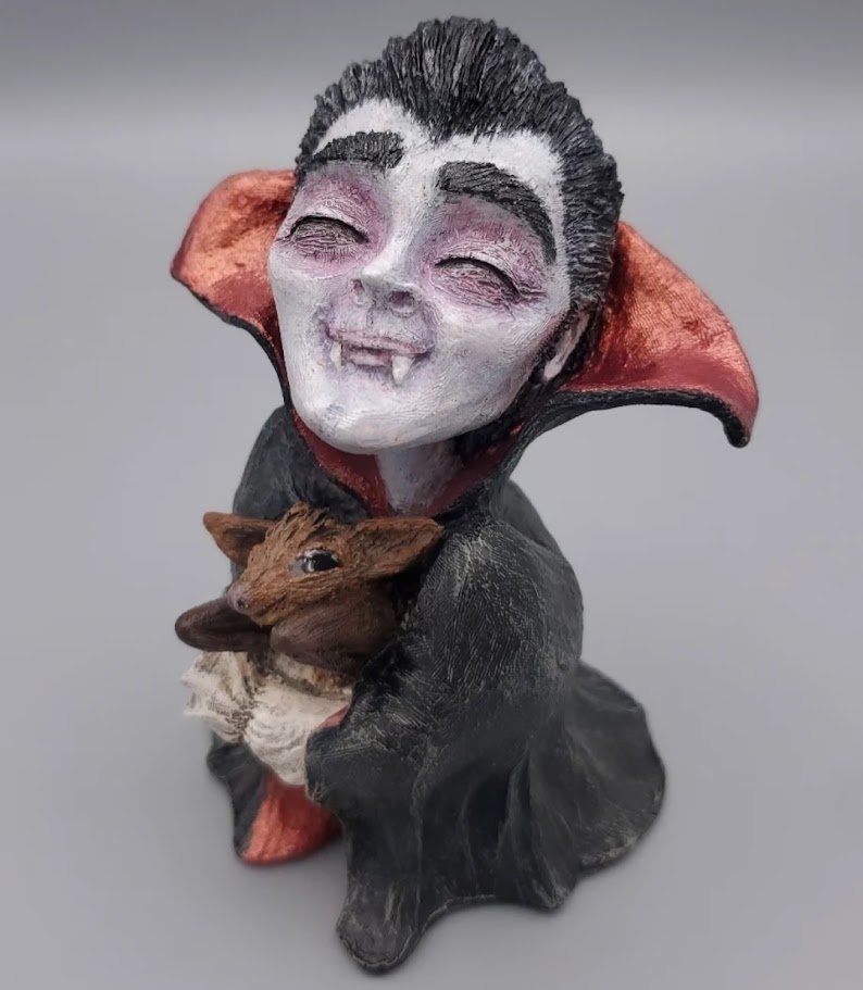 A vampire figurine with a smiling face, fangs, and dark hair, holding a small bat in its arms, against a plain gray background.