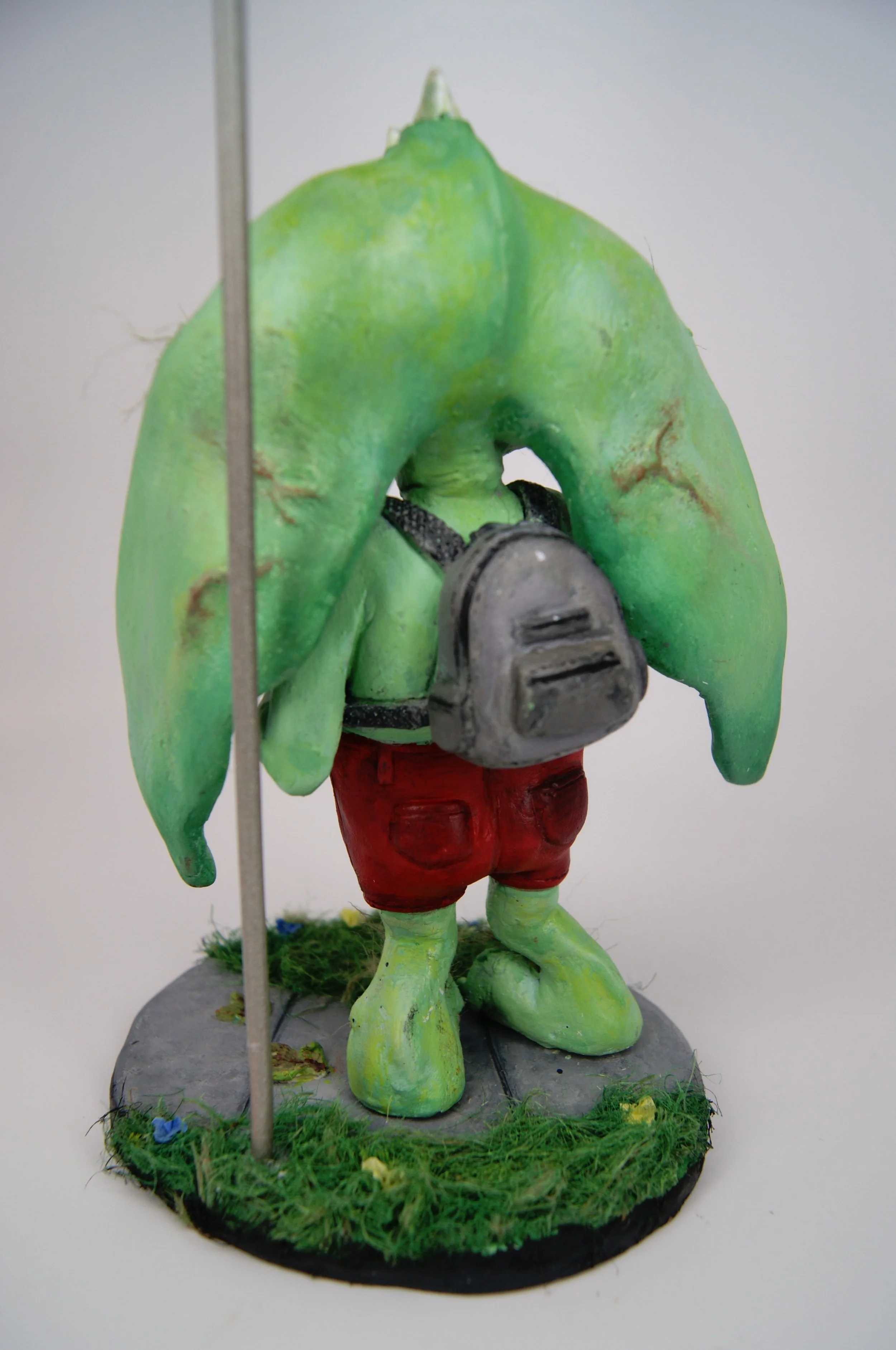 A green anthropomorphic frog figurine wearing red shorts, a backpack, standing on a grassy base.