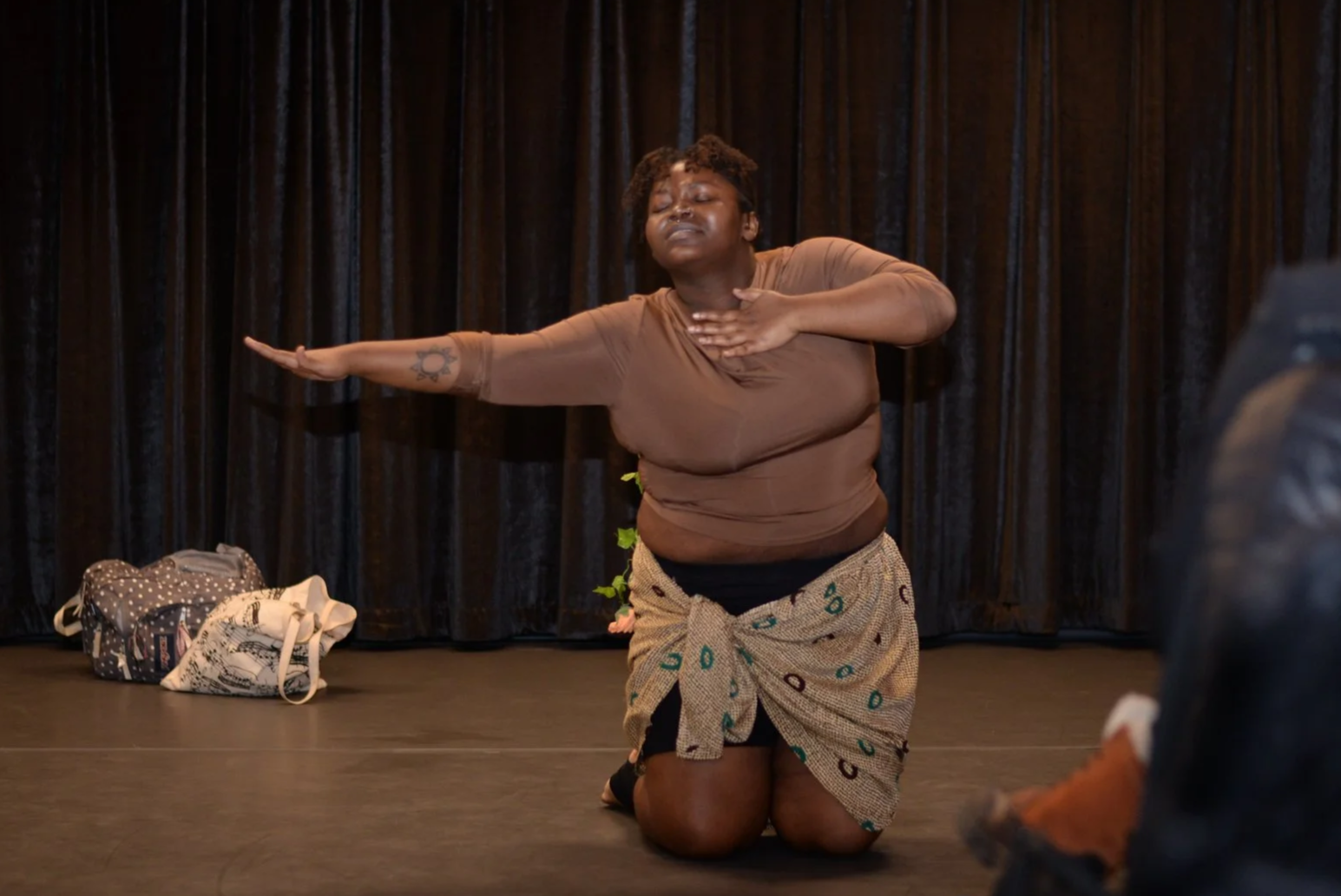 A woman kneels on the stage in front of black curtains, with one arm extended and the other hand on her chest, appearing to be performing or expressing emotion.
