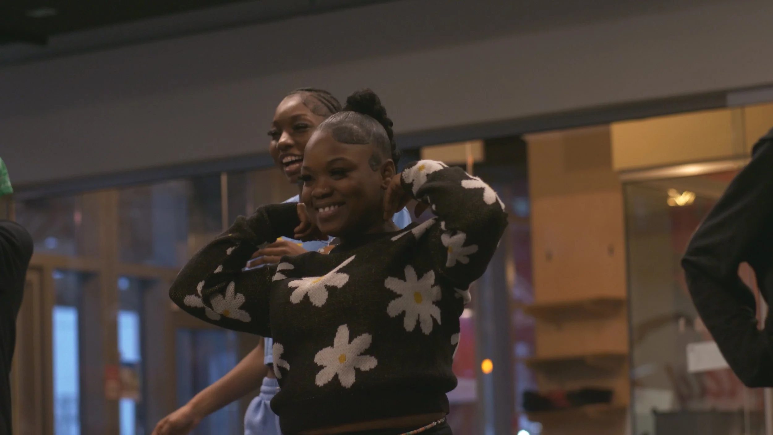 A young girl with a floral sweater smiling and adjusting her hair, standing indoors with a woman behind her.