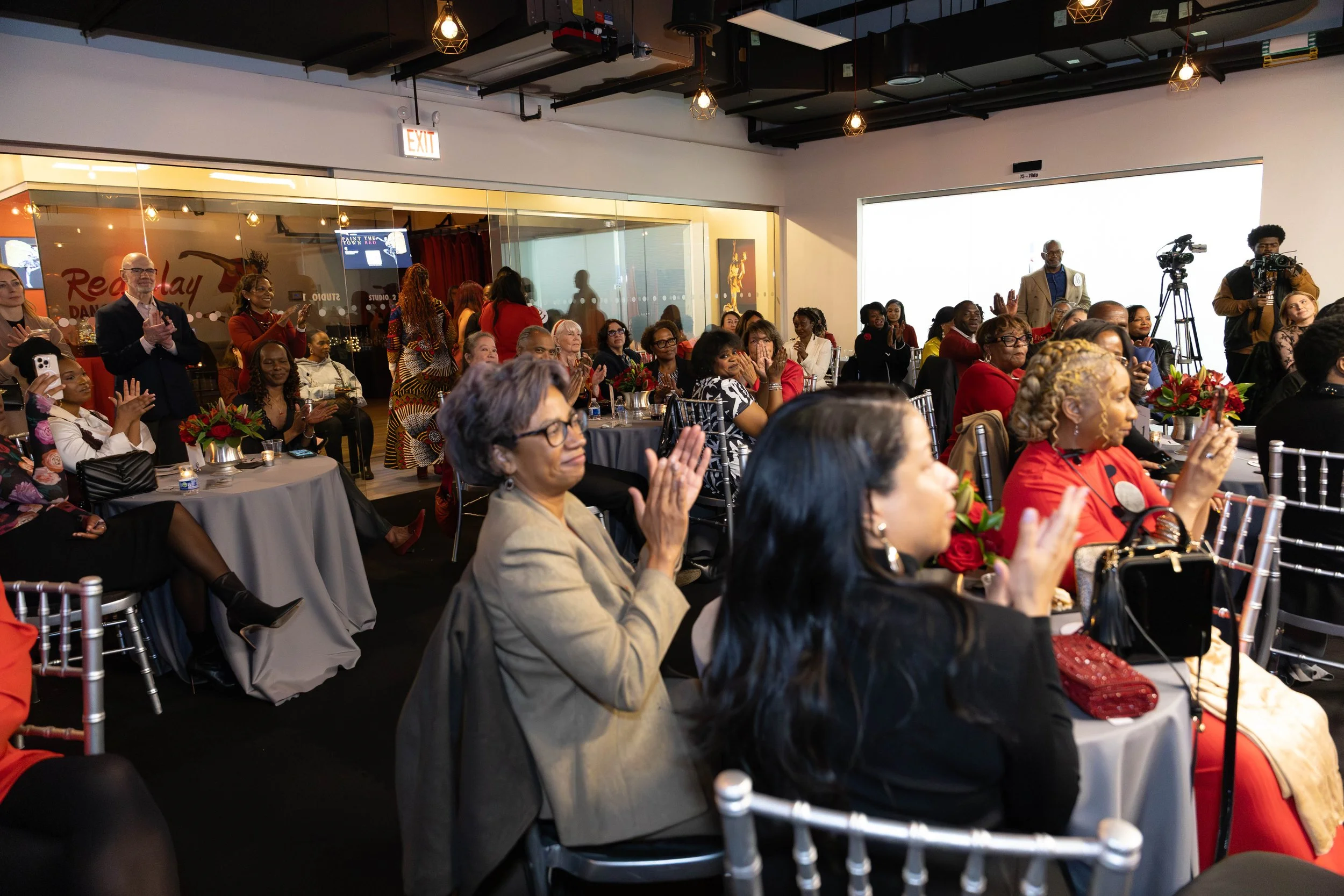 A diverse group of people sitting at round tables, clapping and enjoying an event in an indoor venue with modern lighting and a glass wall.