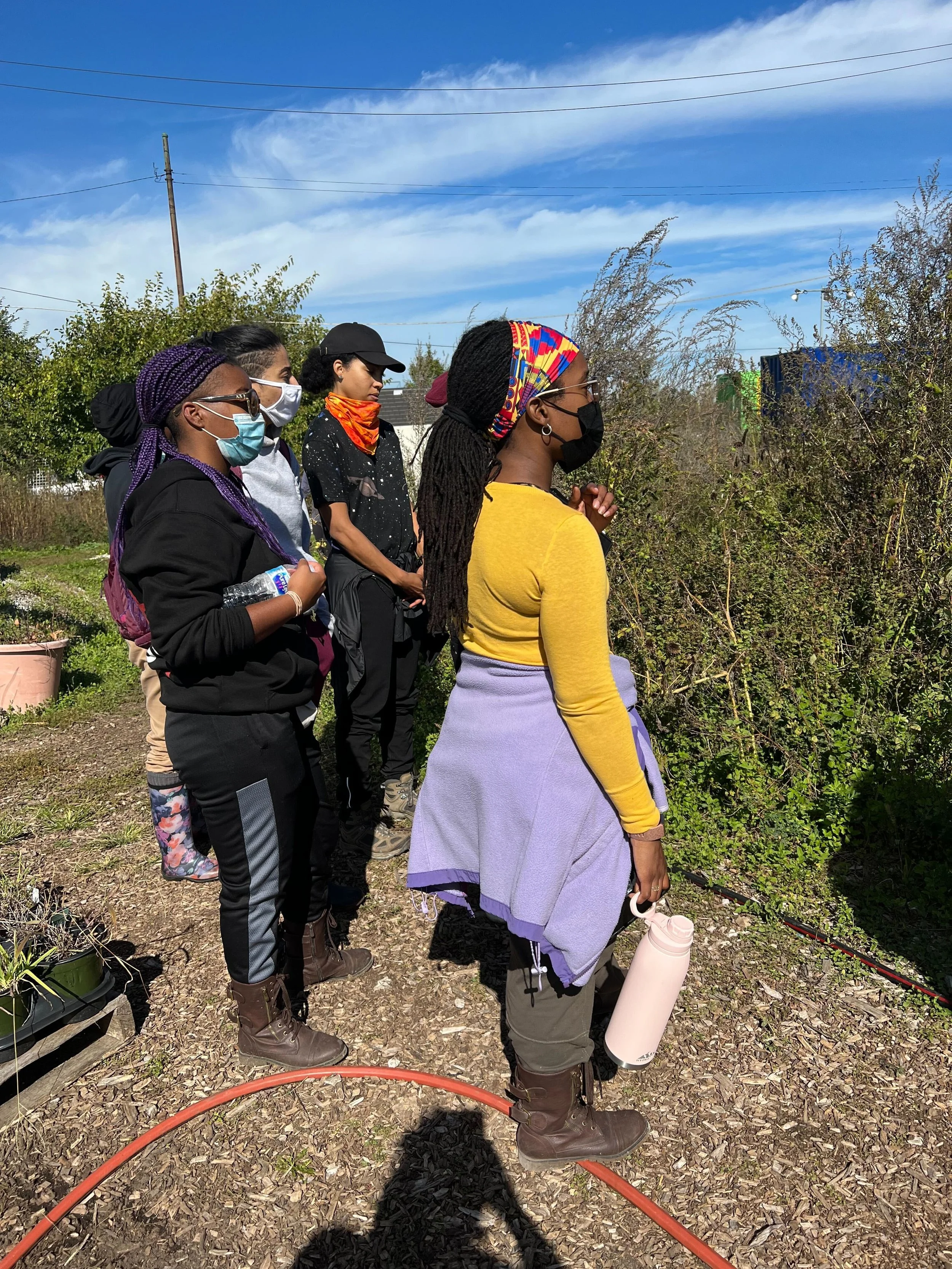 A group of people outdoors wearing masks, standing on a dirt path surrounded by plants, under a blue sky.