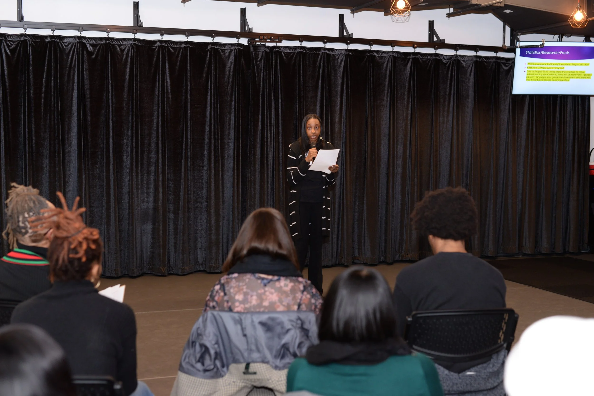 A woman giving a presentation on stage in front of an audience, with a slide projected on a screen to her right. The audience is seated facing the stage, listening attentively.