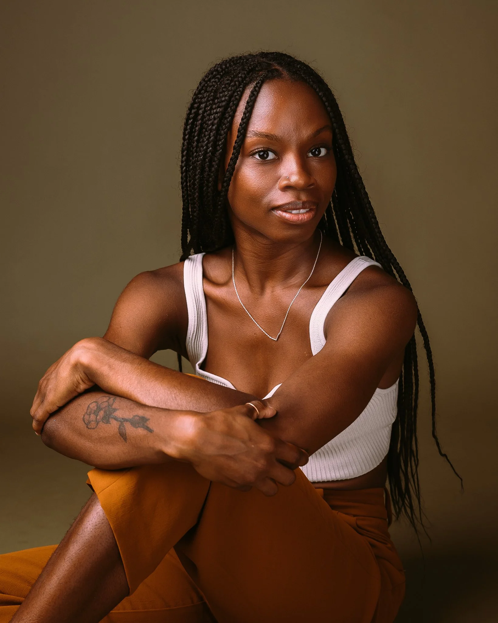 Portrait of a woman with long braided hair wearing a white tank top, copper-colored pants, and a silver necklace, sitting against a plain olive background.