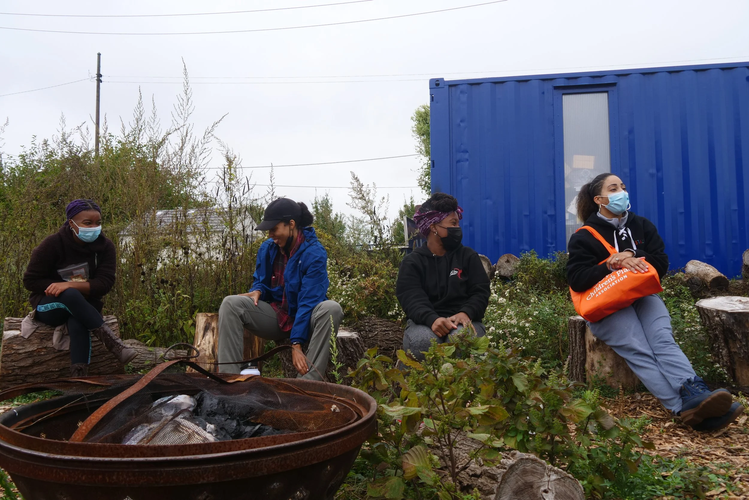 Four people wearing masks sitting around a fire pit outdoors, surrounded by greenery, with a blue shipping container in the background.