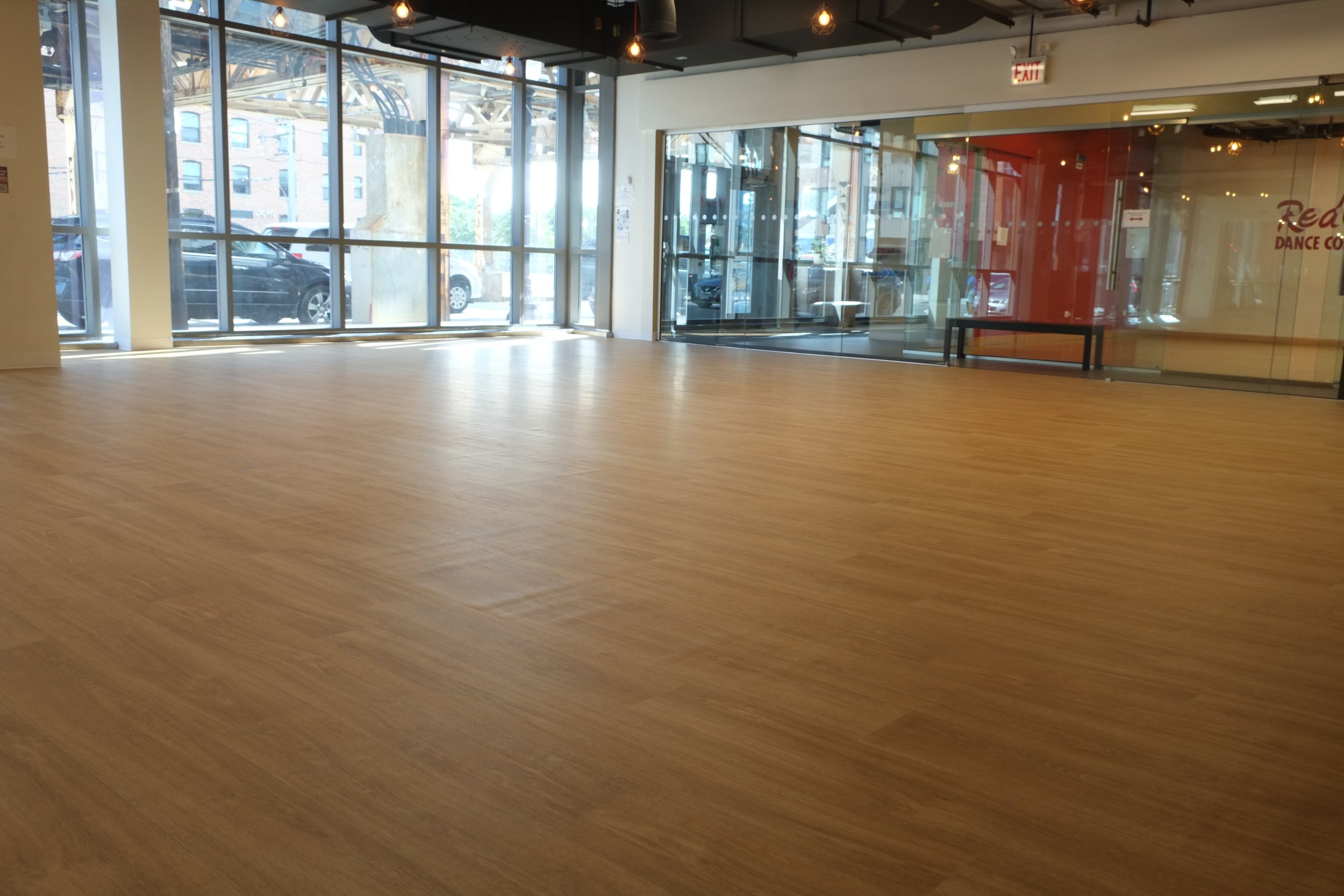 An empty indoor space with large windows and wooden flooring, with a glass door leading outside where parked cars and a building with an elevated structure are visible.