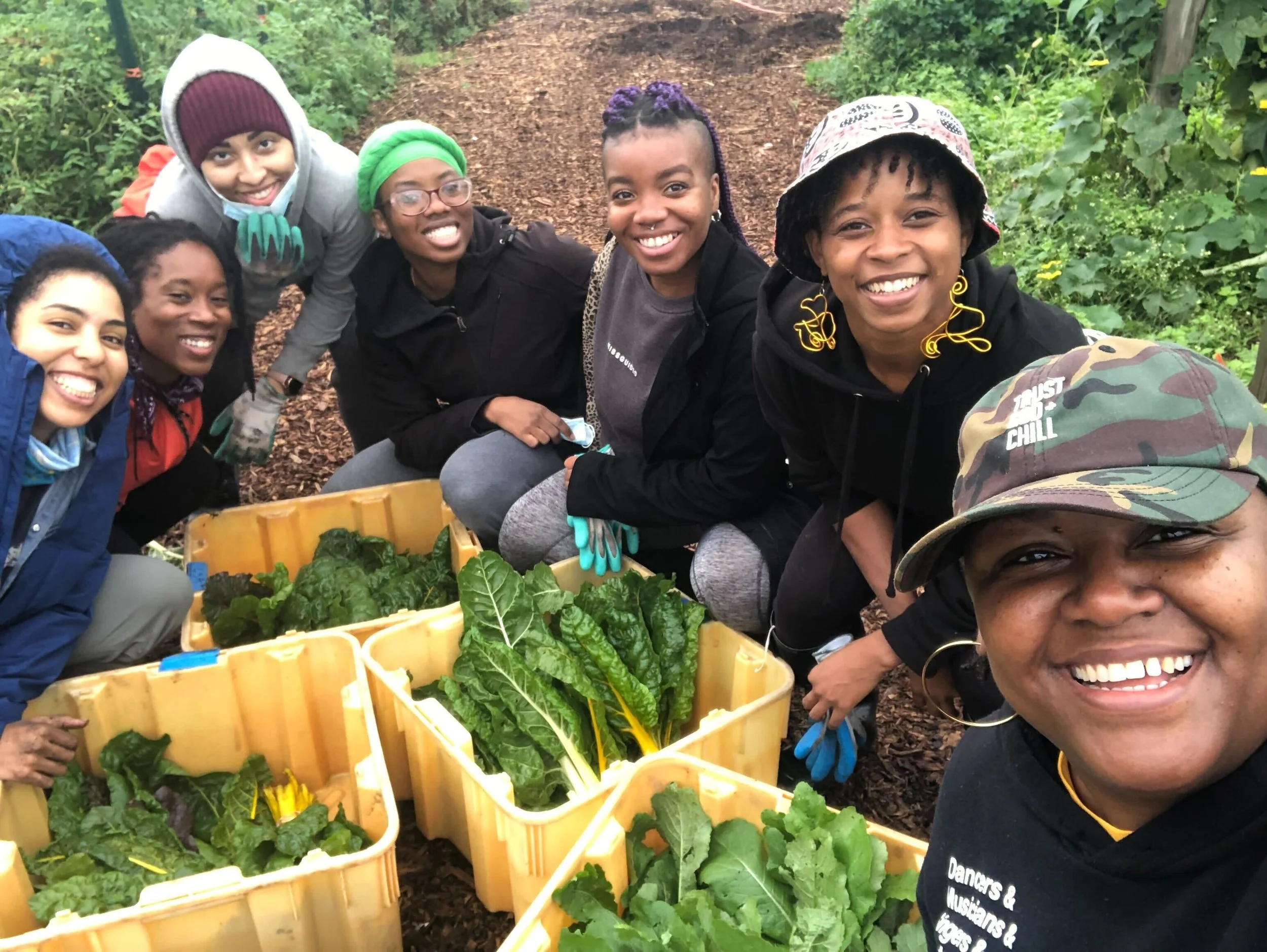 A group of smiling people gather around bins of freshly harvested leafy greens in a garden setting.