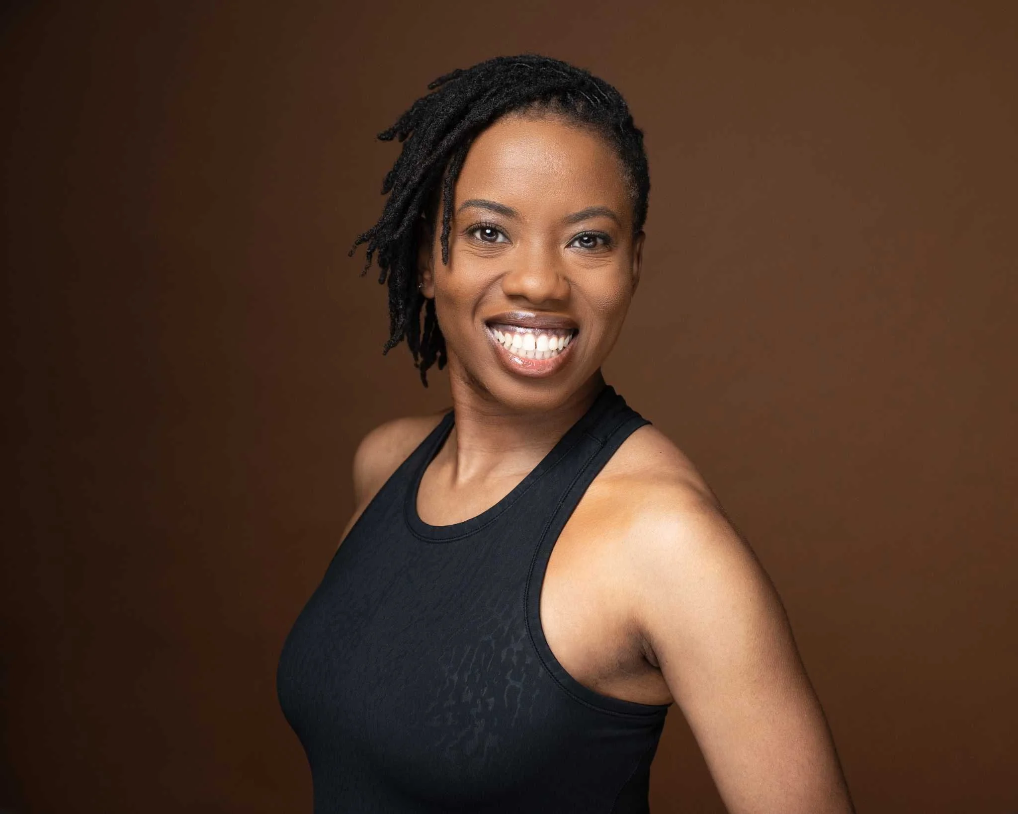 Close-up of an African American woman with short dreadlocks, wearing small earrings, looking directly at the camera with a neutral expression against a plain background.