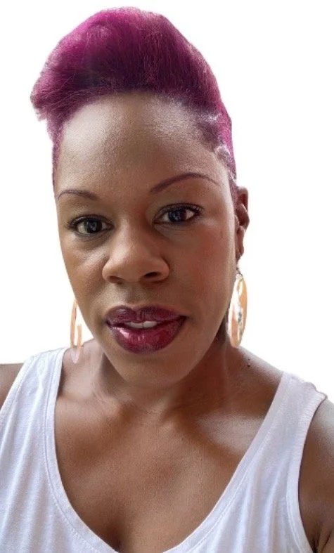 A woman with short, pink hair, large hoop earrings, and wearing a white tank top, looking directly at the camera.