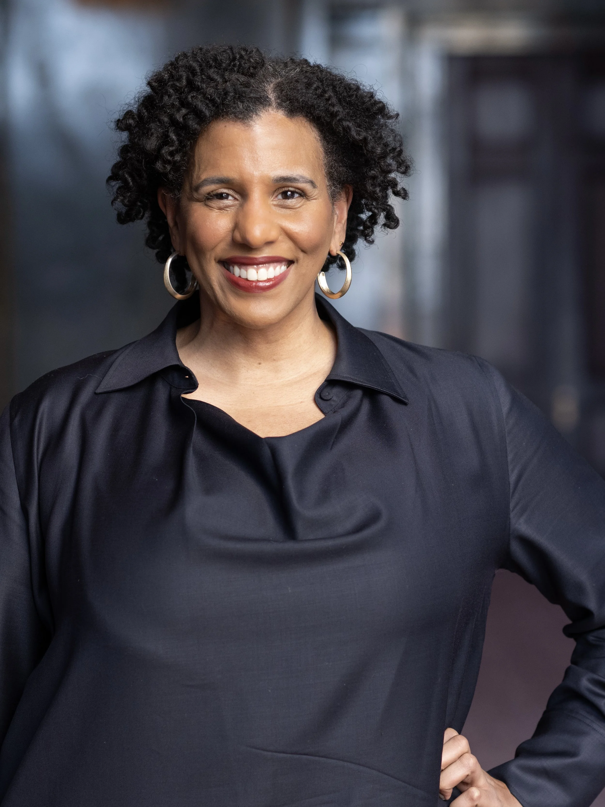 Smiling person with curly hair wearing a dark shirt and hoop earrings.