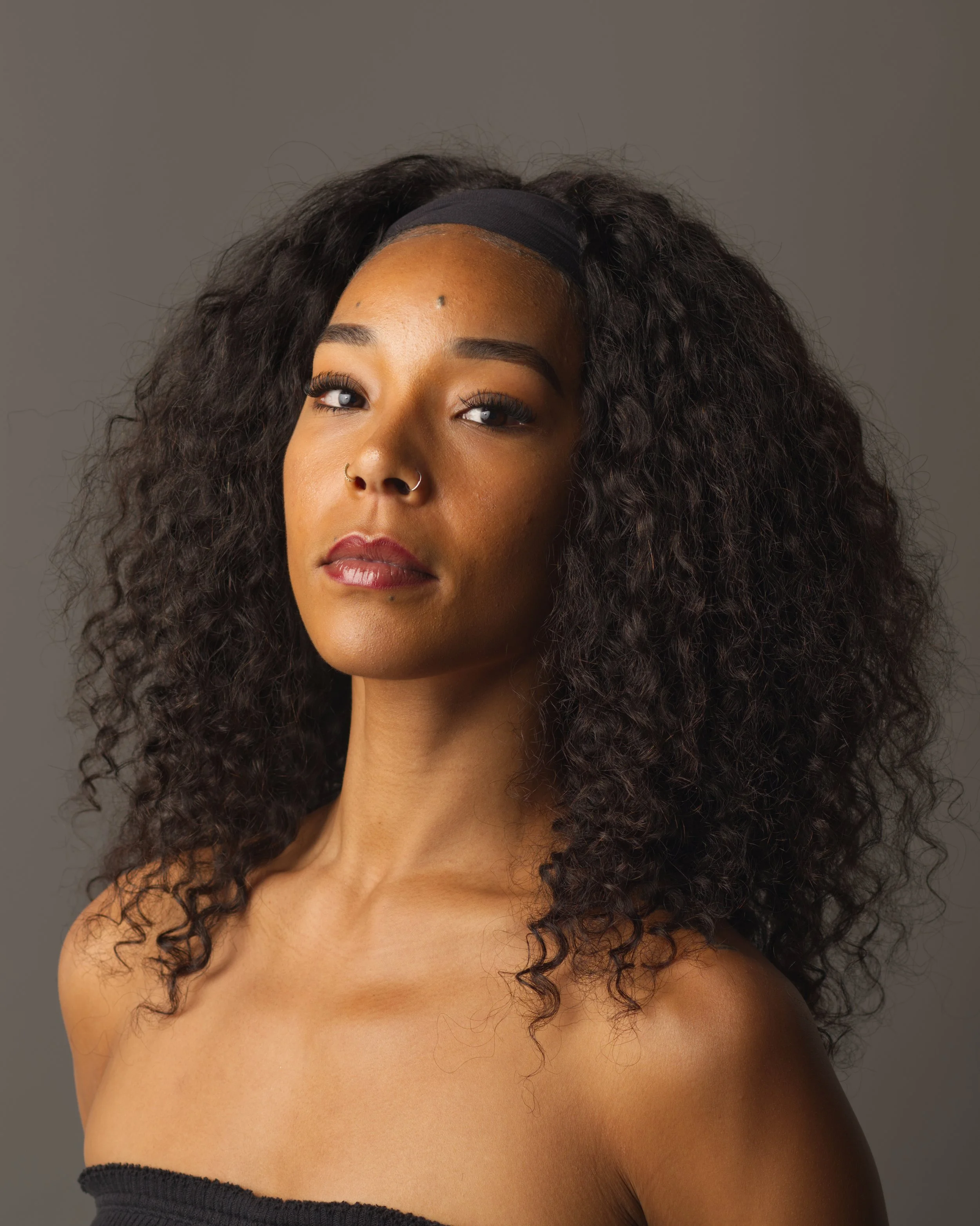 Close-up of a woman with curly hair wearing a black headband, makeup, and a nose ring, with a neutral gray background.