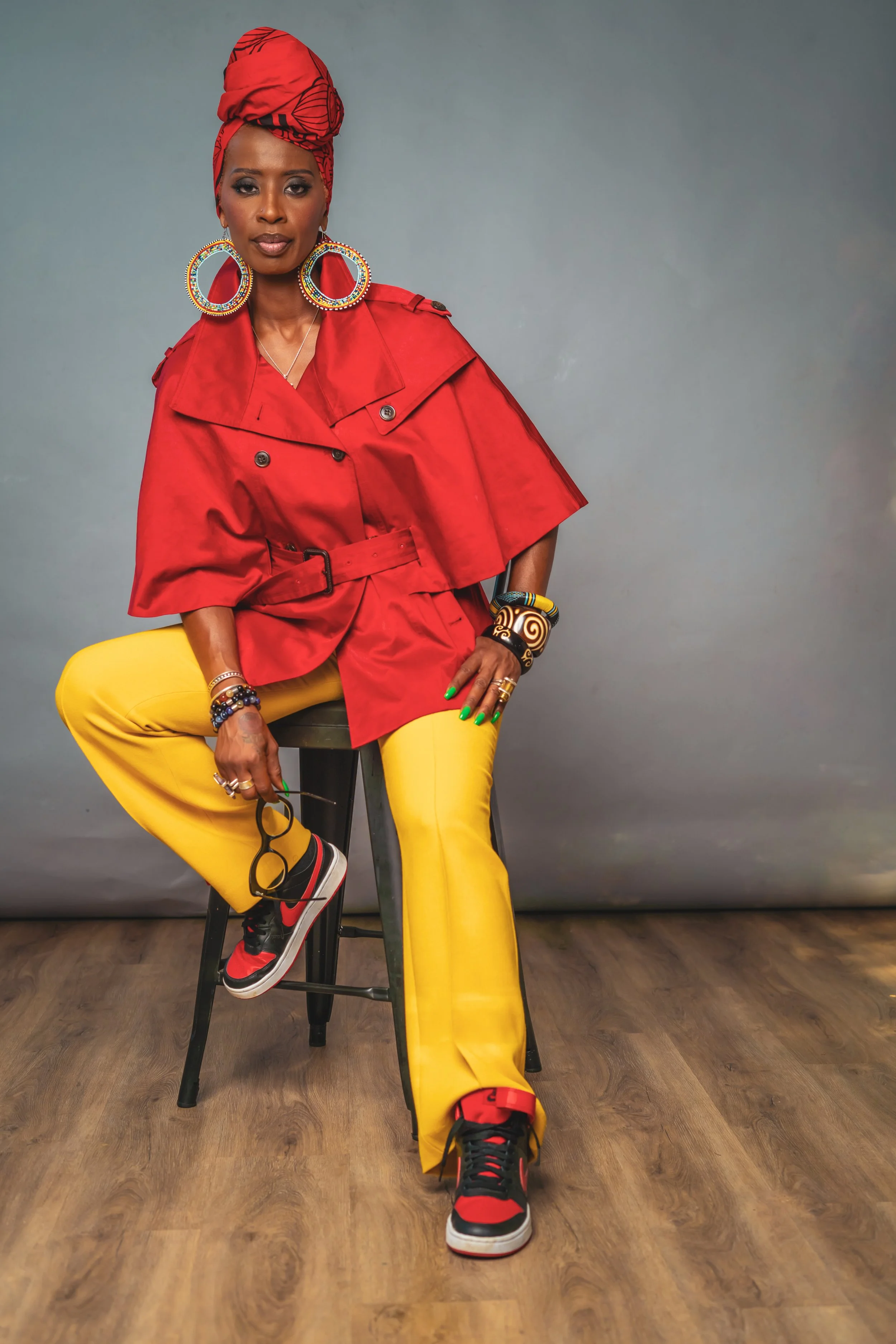Person sitting on a stool wearing a red jacket, red headwrap, large colorful hoop earrings, yellow pants, and black and red sneakers, holding glasses with green nails.