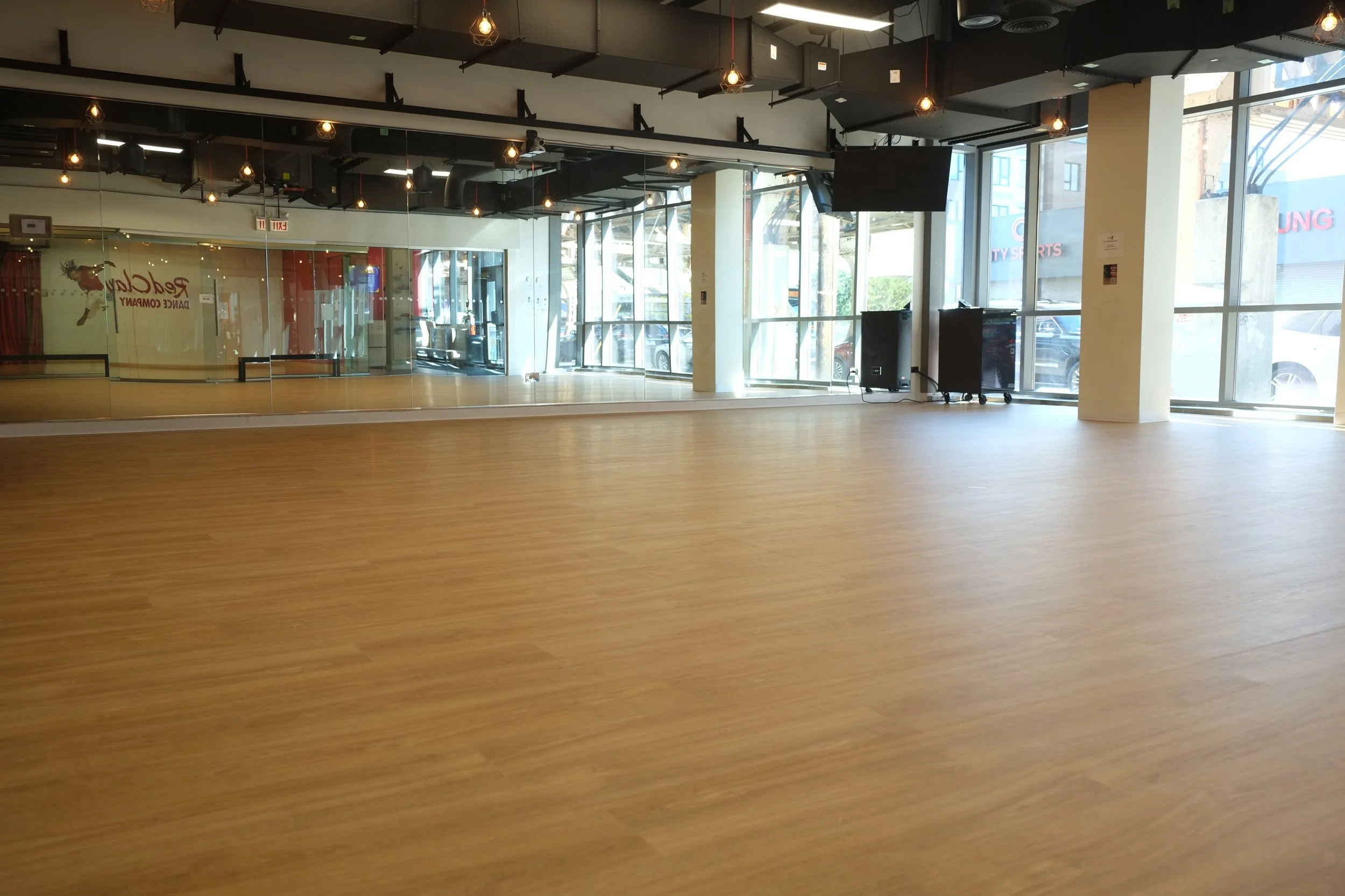 Empty dance studio with wooden floor, large front windows, black ceiling with lighting fixtures, and mirrors on one wall.