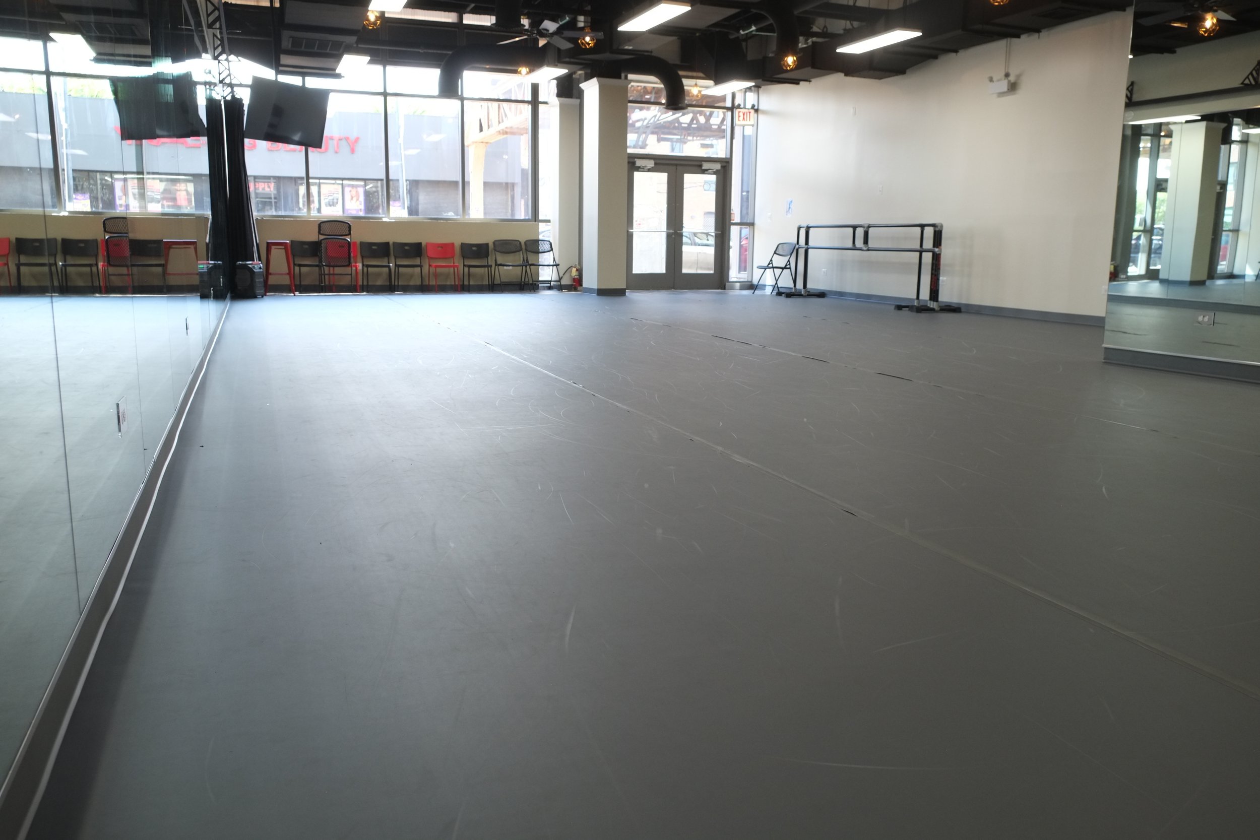 Empty interior space with chairs along a window, a glass door, a large mirror, and a beige wall, suggesting an event or dance studio.