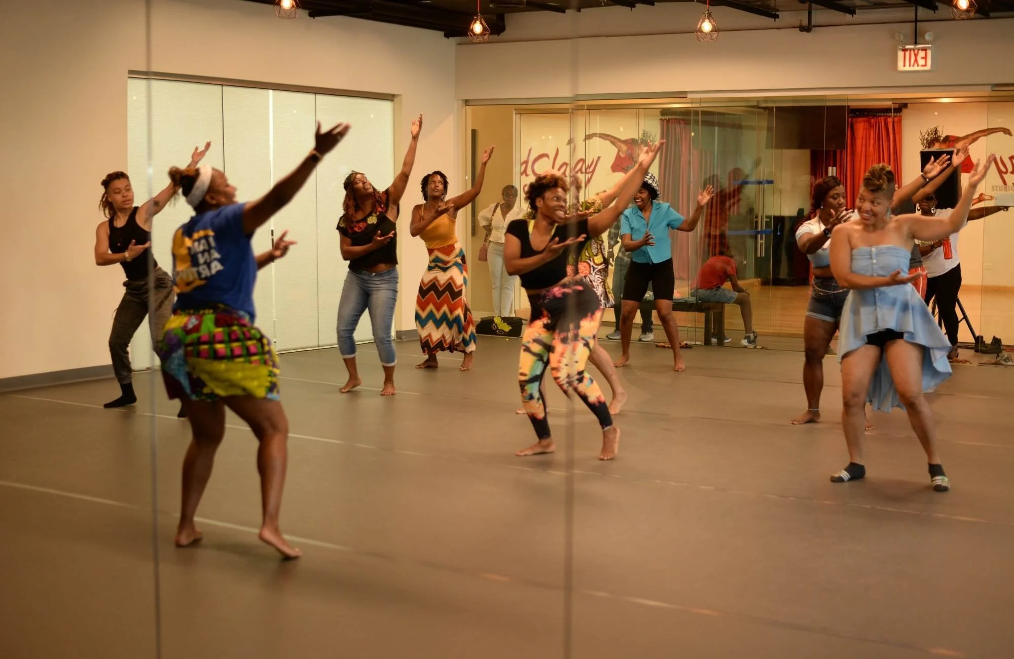 Group of women dancing in a dance studio with a wooden floor and glass wall, some women are barefoot and others wearing socks, while one wears colorful leggings and another a long patterned skirt.