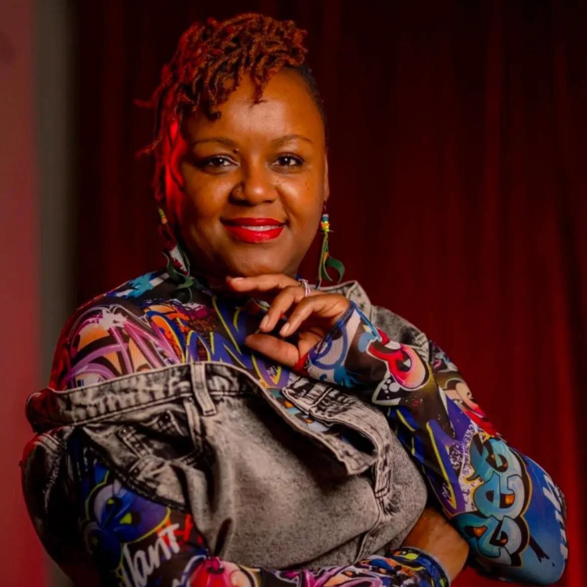 A confident woman with short dreadlocks, red lipstick, colorful earrings, wearing a multicolored graffiti-style shirt and a denim jacket, posing with her hand near her chin against a red curtain background.