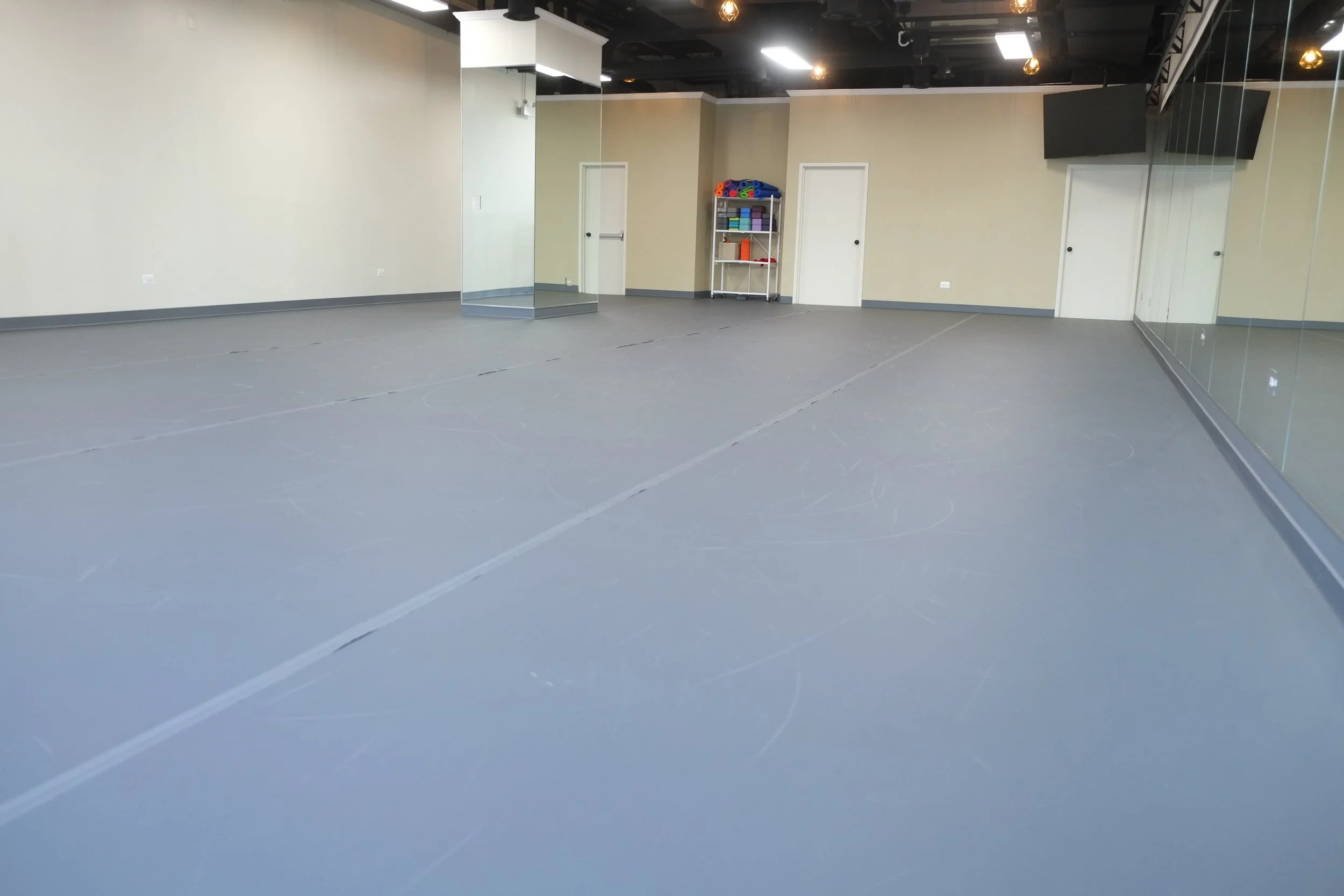 An empty dance or fitness studio with large mirrors on the walls, a small shelf with rolled-up mats and other equipment, and a gray floor with faint tape markings.