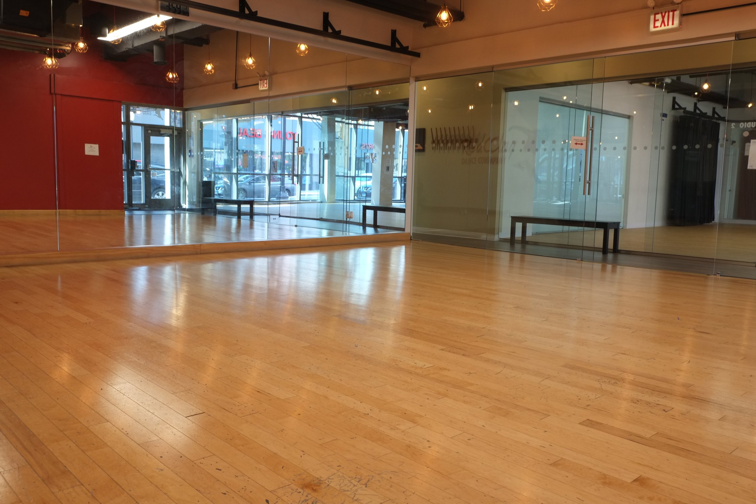 Empty dance studio with large mirror on the wall, wooden floor, and glass doors leading outside. Overhead lights are on, and there is an exit sign above the door.