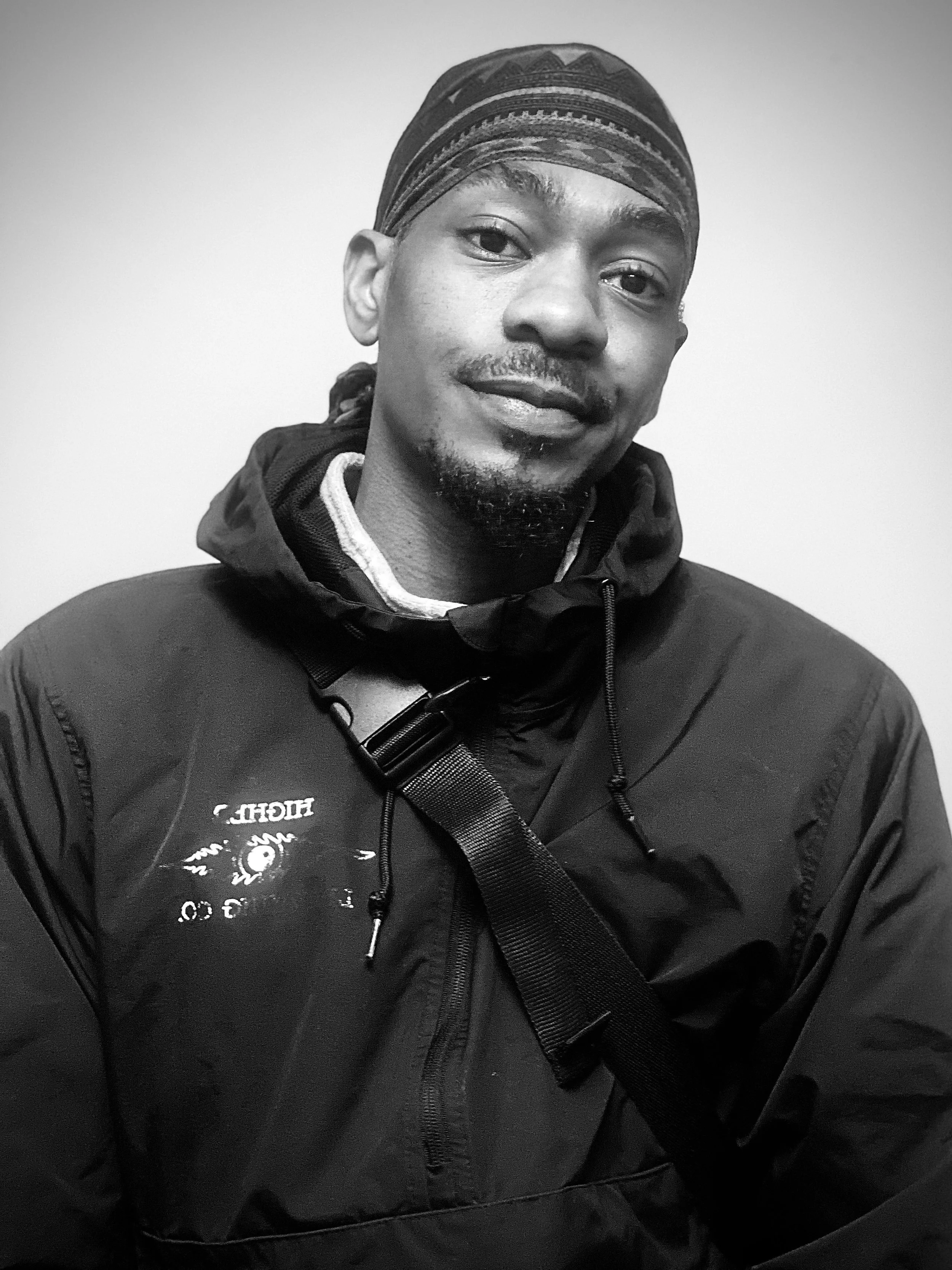 Black and white photo of a young man wearing a cap and a jacket with a strap, smiling slightly, against a plain background.