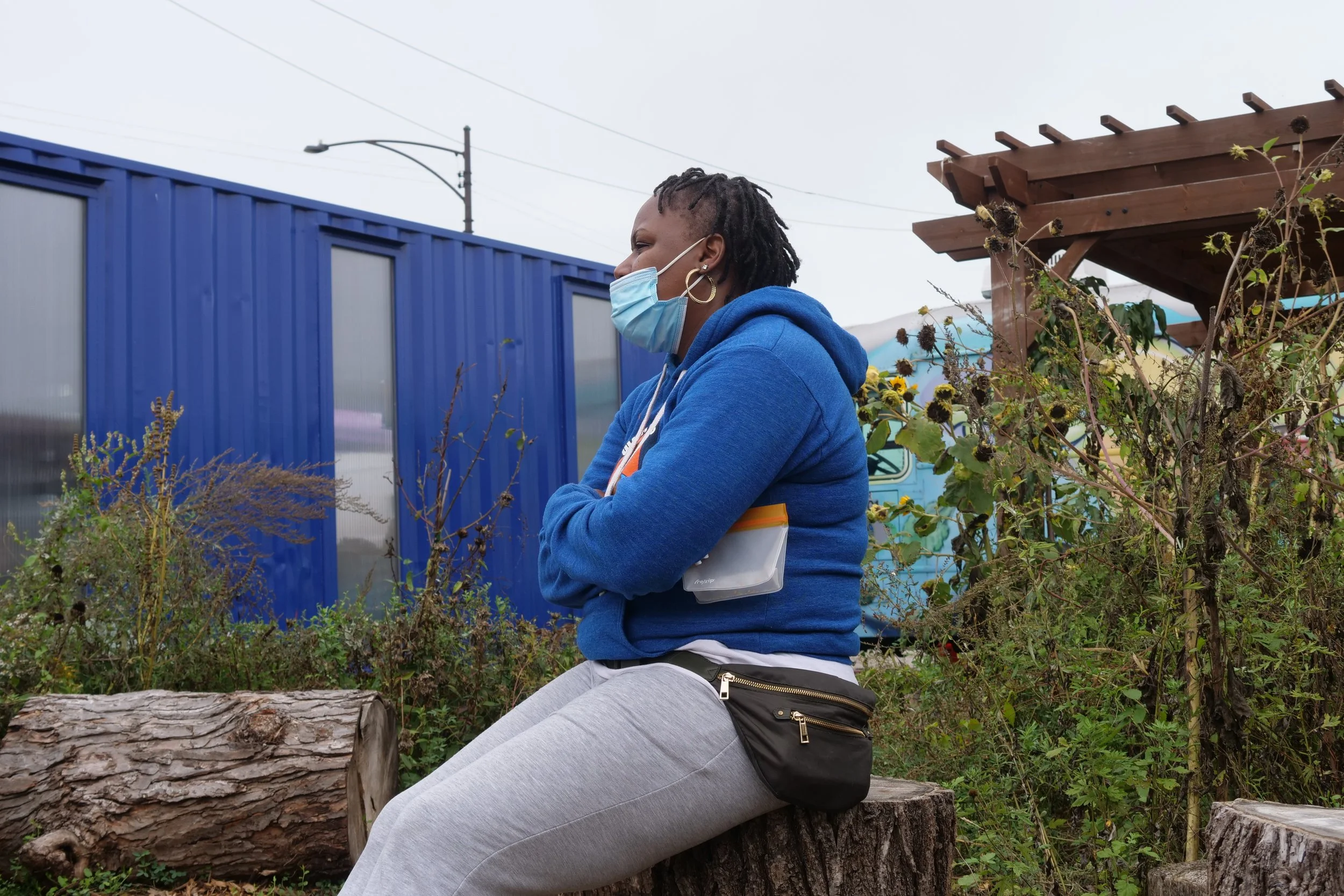 A person in a blue hoodie and face mask sitting on a wooden stump in an outdoor setting with plants, near a blue container and a wooden pergola.