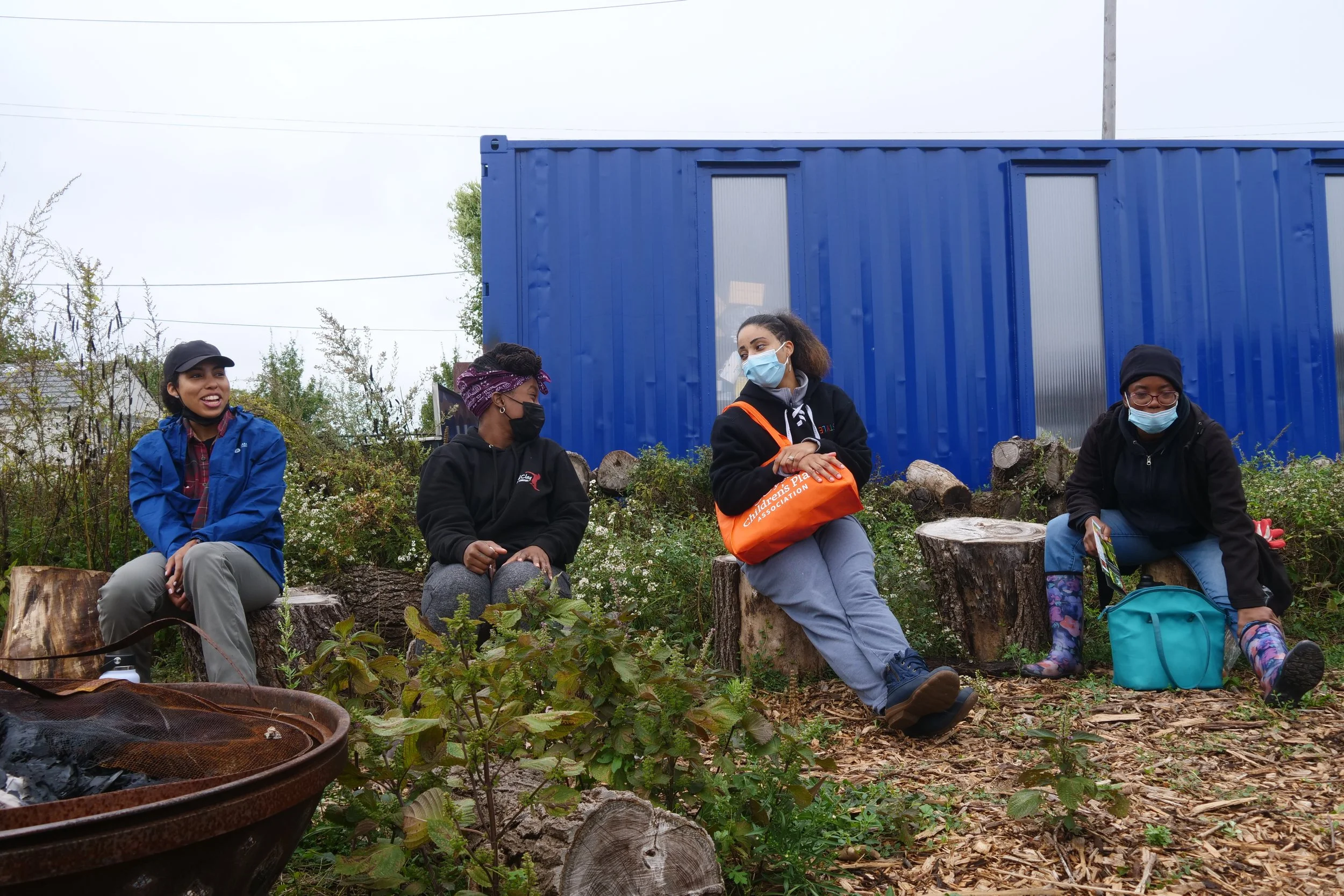 Four people sitting on tree stumps in an outdoor setting with greenery and a blue container building in the background. They are wearing casual clothes, and three are wearing masks.